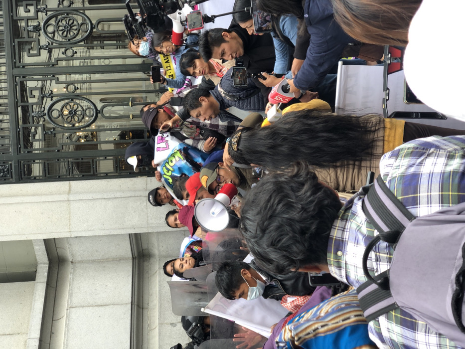 A crowd gathers by a building with ornate metalwork. Some hold phones and a megaphone. Many people wear masks and the group is interspersed with reporters holding microphones. Riot shields are visible near the front of the crowd.