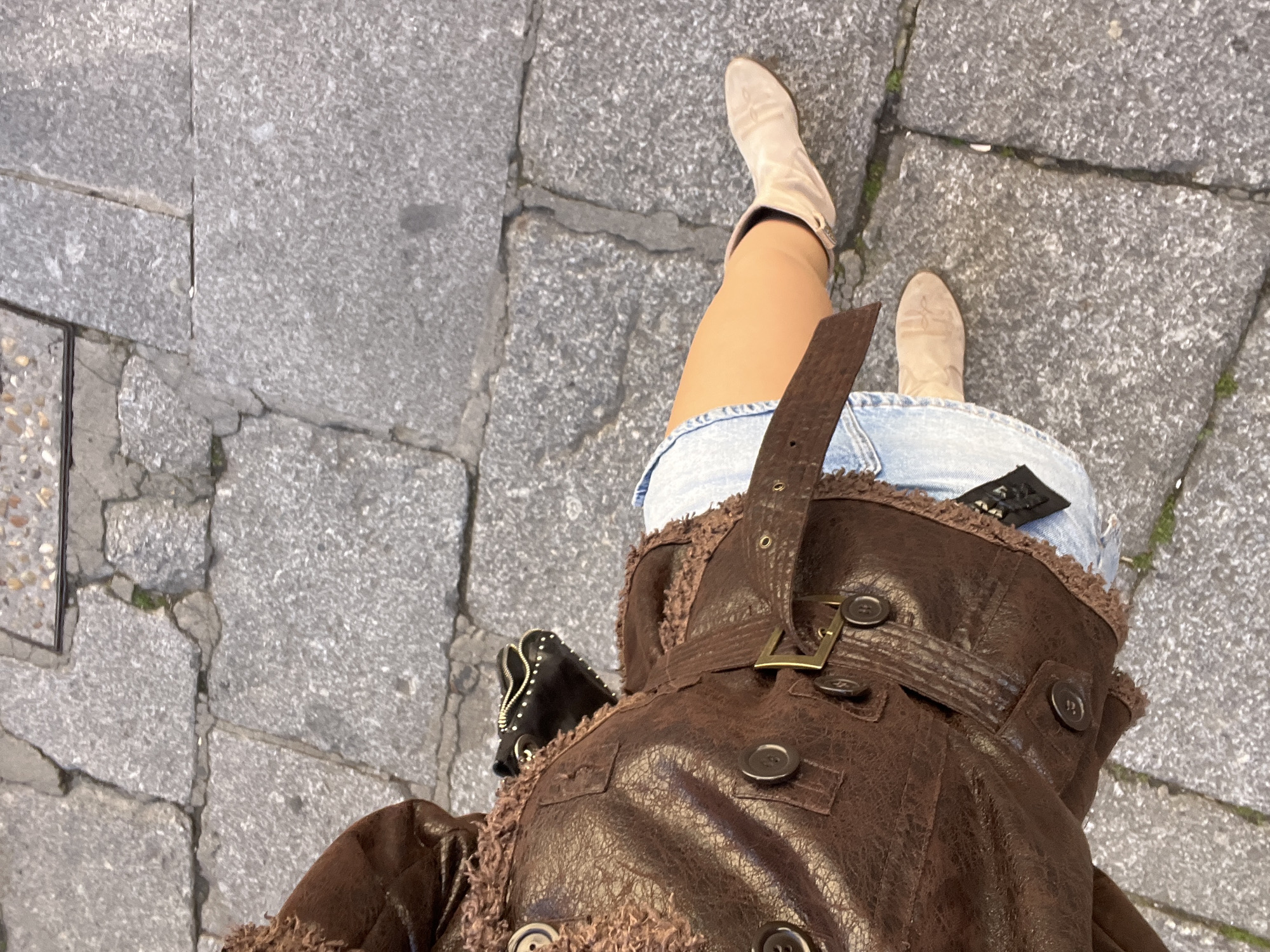 The image shows a downward-angled view of a person standing on a grey stone-paved area in Salamanca, Spain. The paving stones are irregular in shape and size with narrow joints. The person is wearing light beige cowboy boots, a denim skirt, and a brown leather-like jacket with a fur-like trim and a buckled belt. A small black bag with studs is visible on their side.