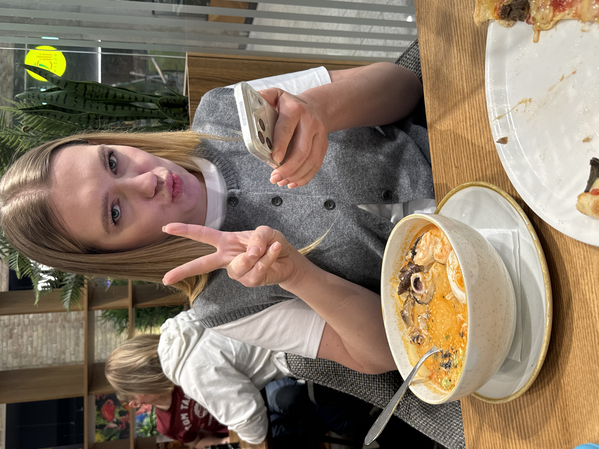 A woman is holding up a peace sign while looking at a phone in her hand in a restaurant. She is wearing a grey sweater over a white shirt. In front of her is a bowl of soup on a plate, as well as a plate with a partially eaten pizza. Another person can be seen in the background, wearing a white jacket and a maroon shirt. The restaurant has wooden shelves with plants and books. A city is not visible. It is not possible to determine if the location is in Riga, Latvia.