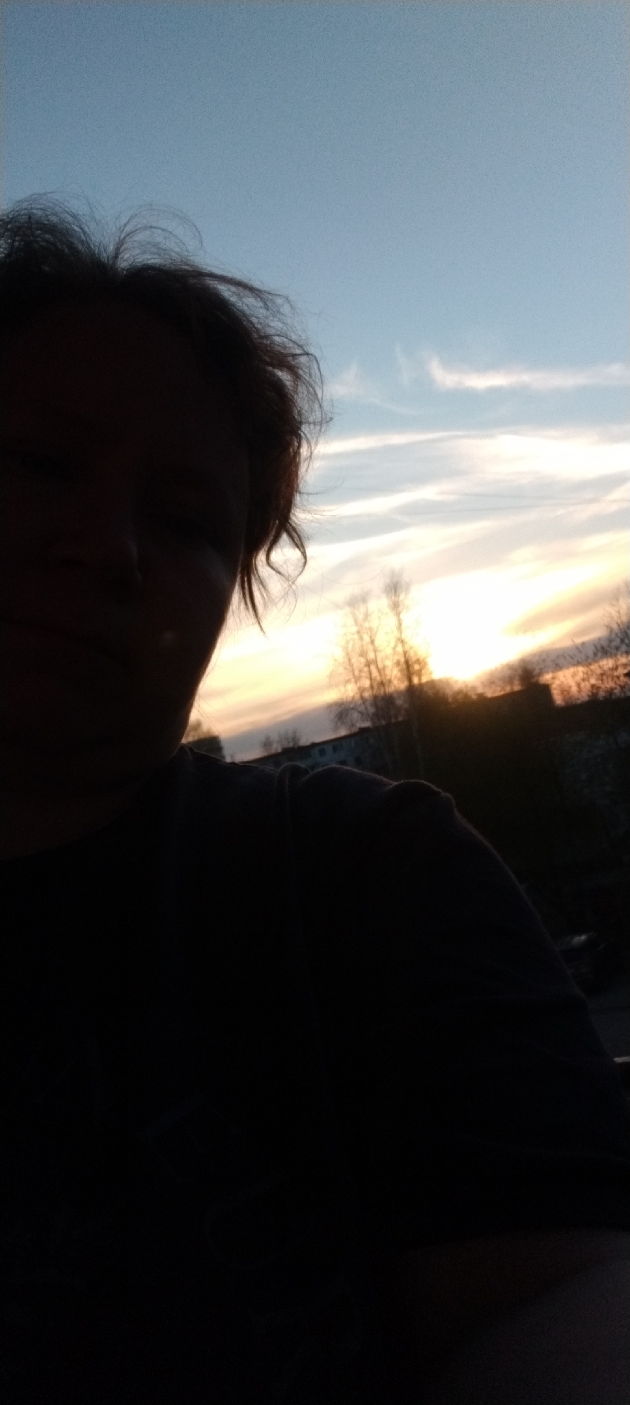 The image depicts a person silhouetted against a sunset sky. The person's face is partially visible in shadow. The background features clouds colored by the setting sun, trees without leaves, and a multi-story building. The location is a city and the sky is visible above.