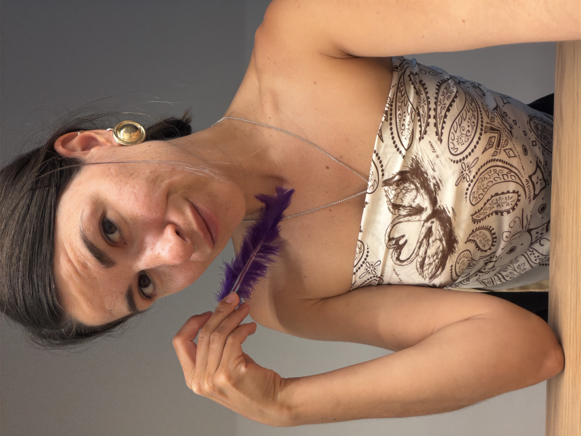 The image displays a close-up portrait of a woman, rotated 90 degrees counter-clockwise. She has dark hair, pulled back, and wears a large, round, gold-colored stud earring on her left ear. Her gaze is directed upwards towards the camera, with a slight, playful smirk on her face. A thin, silver-colored chain necklace rests around her neck.
She is wearing a light-colored top or scarf, possibly wrapped, featuring an intricate brown paisley-like pattern. Faint text, "a time", is visible on the fabric of her top. With her right hand, she holds a vibrant purple feather, gently touching it to her collarbone area. Her arms are visible, showcasing natural skin tones.
The background consists of a plain light gray wall. To her right, a warm-toned wooden surface is partially visible, suggesting a table, railing, or furniture. The lighting appears bright and even, indicating an indoor setting. The overall mood is casual and engaging. The scene is set in Budva, Montenegro.