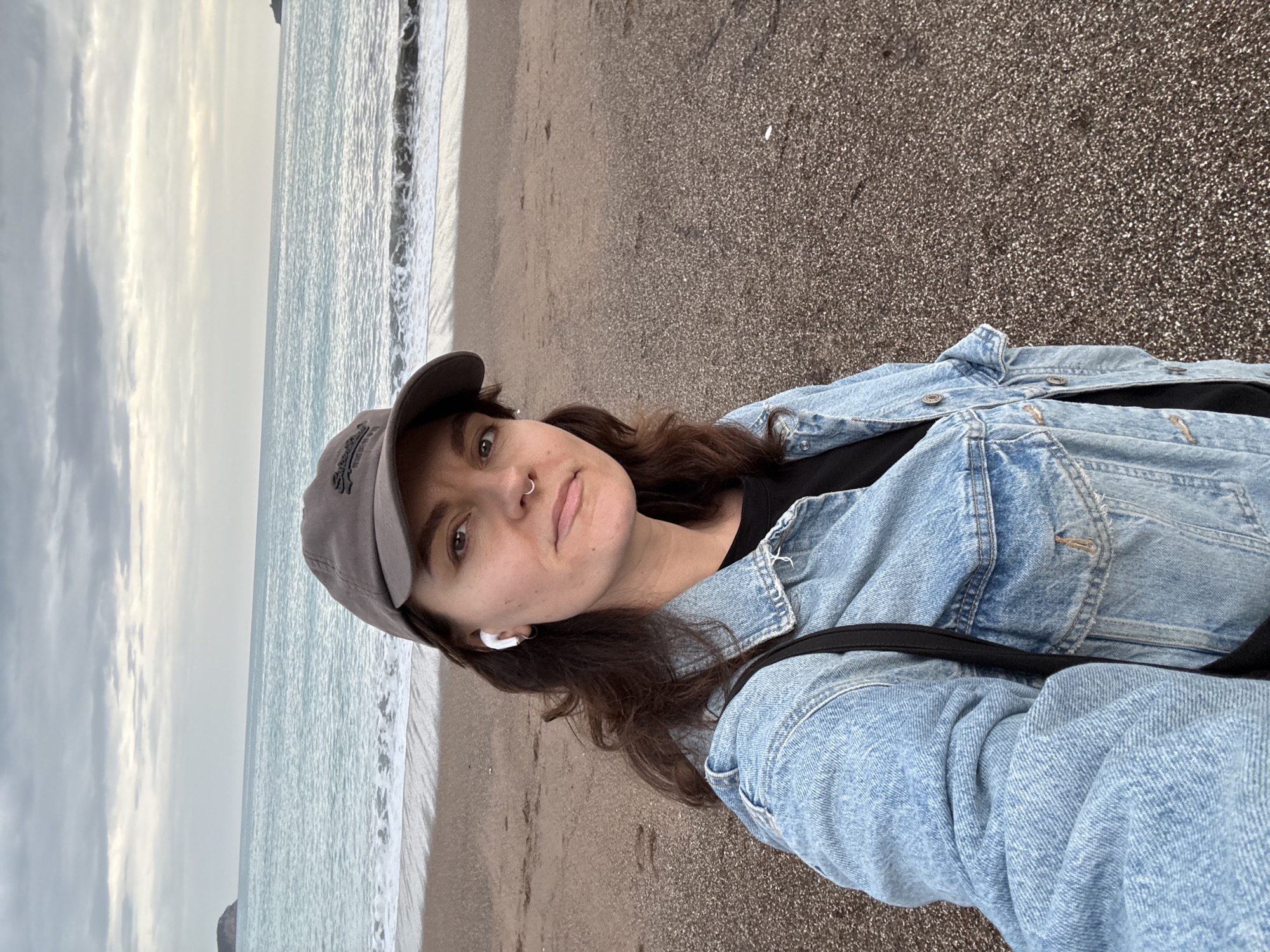 A young woman is standing on a beach in Rafailovići, Montenegro. The beach has dark sand, and the water is light blue with white waves. The sky is cloudy and overcast. The woman is wearing a gray baseball cap, a denim jacket, a black t-shirt, and white earbuds. She has brown hair and a nose ring. A black strap from a bag is visible across her torso. She is taking a selfie.