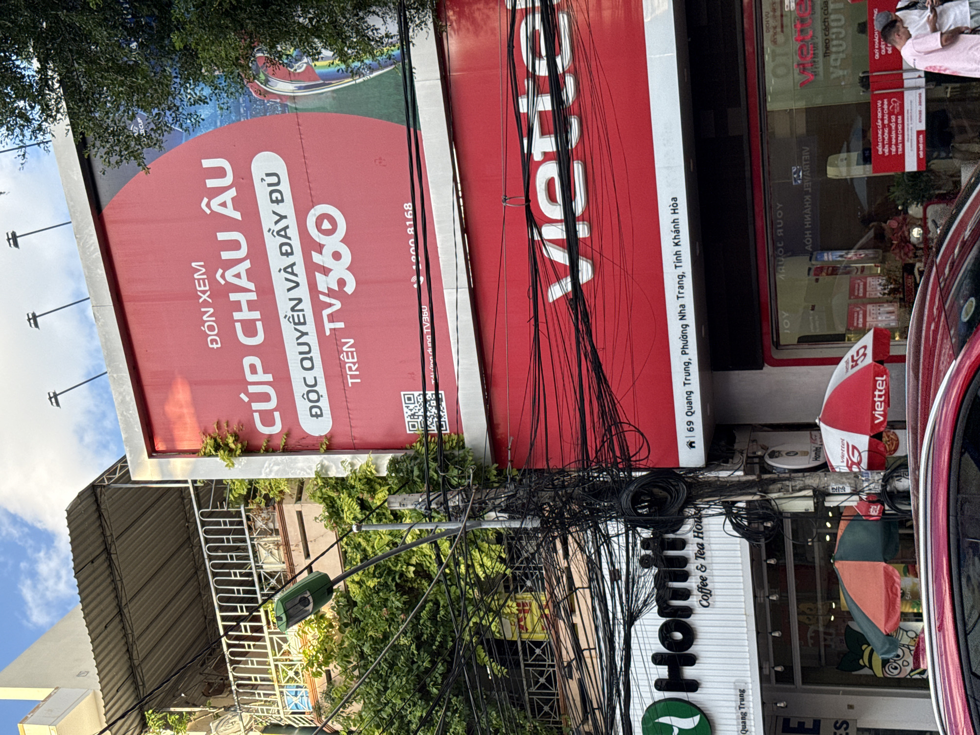 The image captures a street scene in Nha Trang, Vietnam, focusing on commercial buildings and advertisements. Prominently displayed are two large billboards and storefronts. The upper billboard, a vibrant red, features Vietnamese text advertising "ĐÓN XEM CÚP CHÂU ÂU" (Watch the European Cup) and "ĐỘC QUYỀN VÀ ĐẦY ĐỦ TRÊN TV360" (Exclusive and Full on TV360). Below this, another red billboard displays the "Viettel" logo, a telecommunications company. The storefronts below the billboards show signage for "Homita Coffee & Tea House" and another establishment with Vietnamese text. The area is characterized by a dense network of overhead electrical wires, suggesting an urban environment. Several umbrellas are visible, two of which are branded with "Viettel." In the foreground, the top of a red car is visible, and two individuals are standing near the storefronts, one wearing a pink shirt and the other a white shirt. The overall impression is a busy commercial street with a strong presence of advertising and infrastructure.