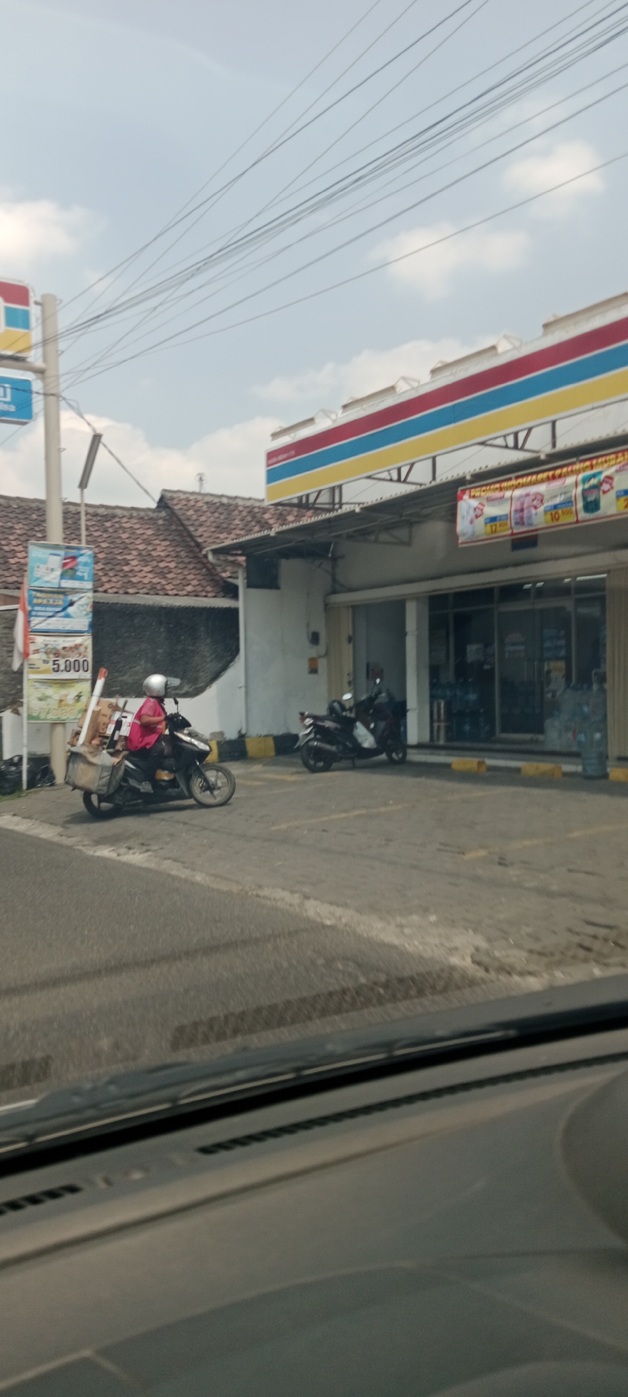A daytime, eye-level, medium-wide shot captures a street scene outside a convenience store in Nogotirto, Indonesia. The foreground shows the hood and windshield of a vehicle. In the middle ground, a person on a scooter wearing a pink shirt and white helmet is seen from the side, with boxes and a large bag strapped to the back of the scooter. The scooter is in motion, traveling on a gray asphalt road with visible yellow and white painted lines. To the right, another scooter is parked in front of a building. The building is a convenience store with a white facade, a striped awning in red, blue, and yellow, and large glass doors. Advertisements are displayed on the store. To the left of the store, a pole with signs and a national flag is visible, along with a building with a tiled roof. The sky is light blue with scattered clouds, and power lines are strung across the upper portion of the image.