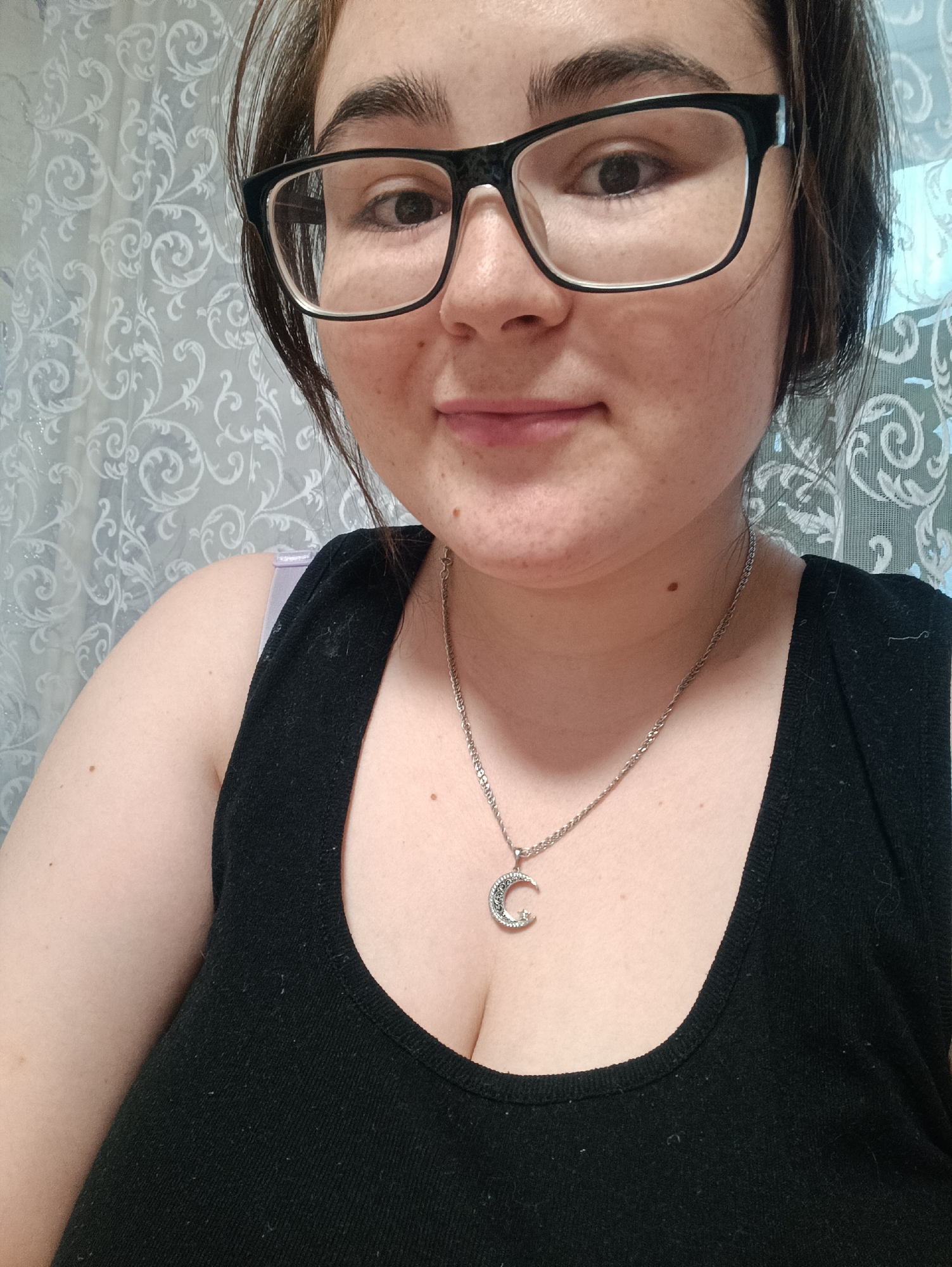 A woman with fair skin, wearing black-framed glasses, a black tank top, and a silver necklace with a crescent moon pendant is shown in a close-up self-portrait. She has dark brown hair and is looking directly at the camera with a neutral expression. In the background is a white curtain with a floral pattern. No specific details indicate this image was taken in Noviy, Russia.