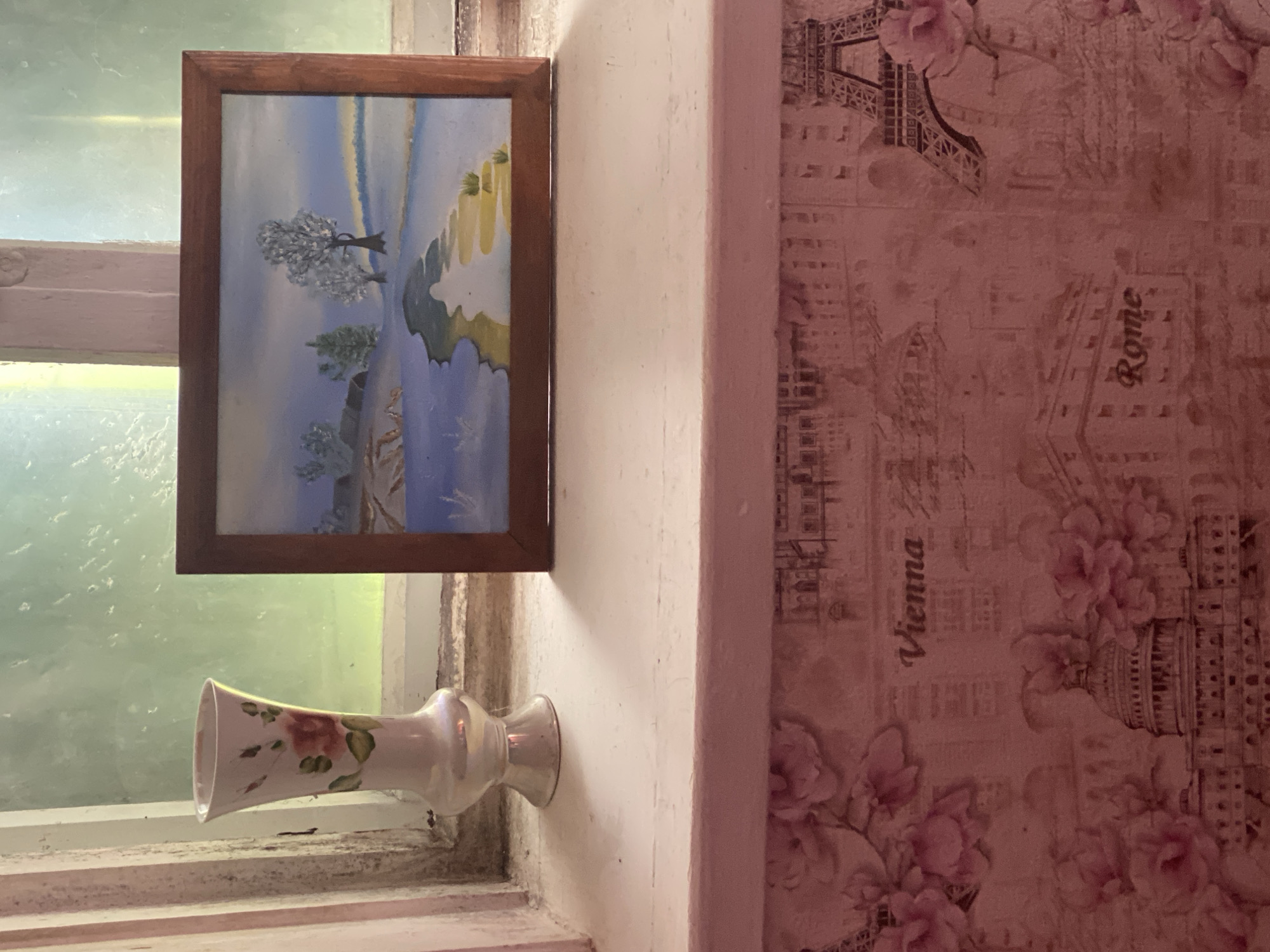 A close-up shot captures a scene indoors, possibly in Ustyvytsia, Ukraine, exhibiting a blend of personal and architectural details. A painting in a wooden frame hangs on a light-colored wall adjacent to a window. The painting depicts a landscape with stylized trees and land formations against a blue sky. Below the painting, a white ceramic vase with floral details sits on the windowsill. To the right of the painting, the wall transitions into a different aesthetic, featuring wallpaper with illustrations of famous cities like Rome and Vienna, interwoven with pink floral designs. The overall lighting and color palette suggest an intimate, personal space.