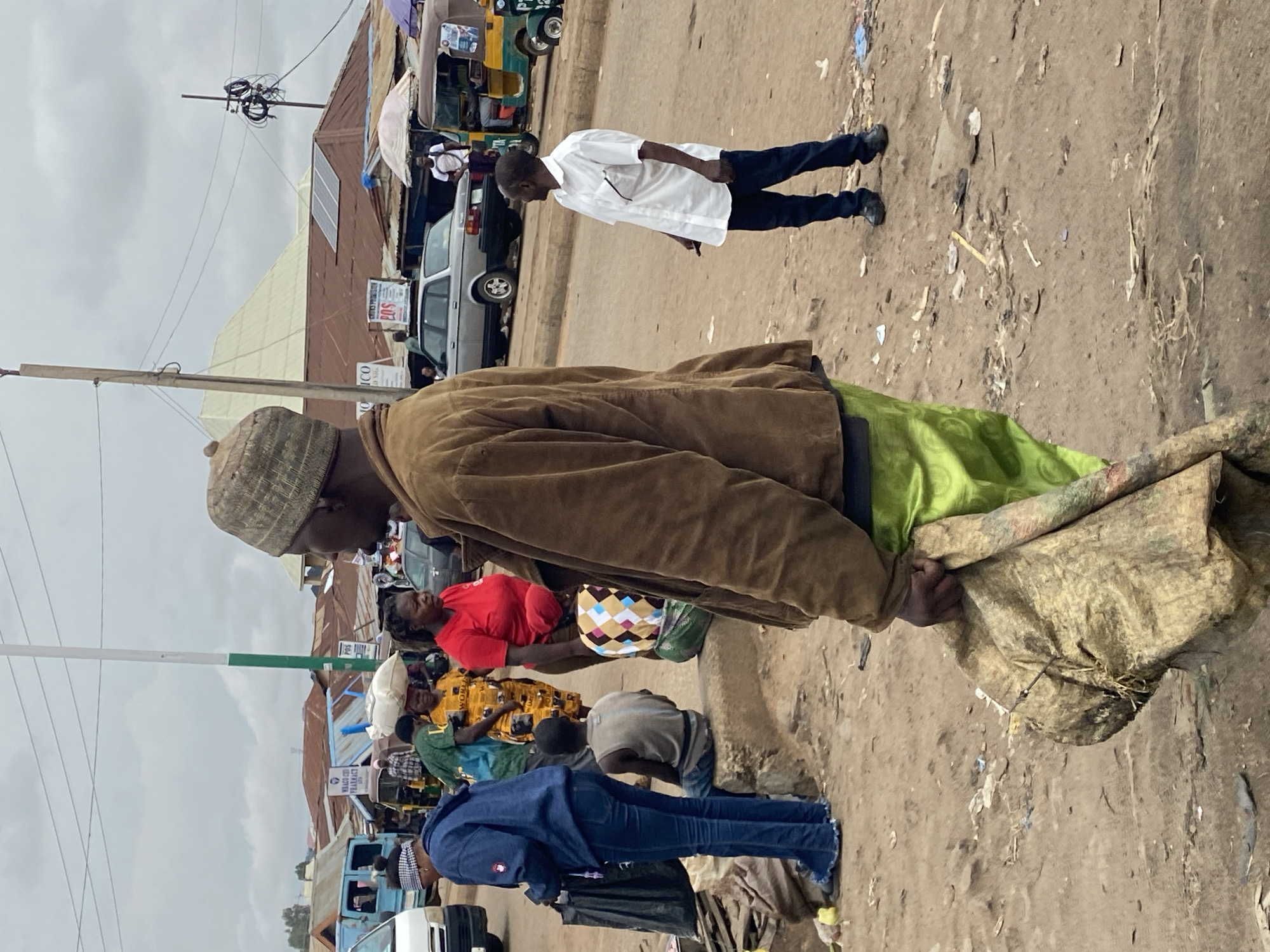 This image captures a bustling street scene in Bukuru, Nigeria, on an overcast day. The ground is unpaved, appearing as dirt or sand, scattered with litter and debris.
In the foreground, a man wearing a brown jacket and a textured, possibly knitted, cap is prominently featured. He is carrying a large, light green and beige sack over his shoulder. To his right, other individuals are visible: a person in a blue jacket and jeans, a woman in a bright red top and a patterned skirt, and another person in a yellow and black patterned shirt. A person is also seen bending down towards the ground.
In the midground, the street is active with several people. A man in a white long-sleeved shirt and dark trousers stands looking towards the left side of the frame. Various vehicles are present, including a silver sedan, a blue minibus, and parts of tricycle-like vehicles (likely 'keke napep'). Buildings line the street, many featuring corrugated iron roofs. A notable detail is a building with solar panels installed on its roof, suggesting an alternative energy source. Utility poles with a dense tangle of wires are also visible against the sky.
The scene depicts everyday life in an urban or semi-urban setting. People appear to be walking, standing, and potentially engaging in commerce, though no explicit transactions are visible. The atmosphere suggests a typical busy day.
Visible text on signs includes "CENTURY PROGRES..." and "EPS," "ICO," and what appears to be "WARCH PHARMACY" and "PBS" on various commercial signs along the street. The sky is uniformly cloudy, indicating an overcast condition, possibly early morning or late afternoon, or simply a consistently cloudy day.