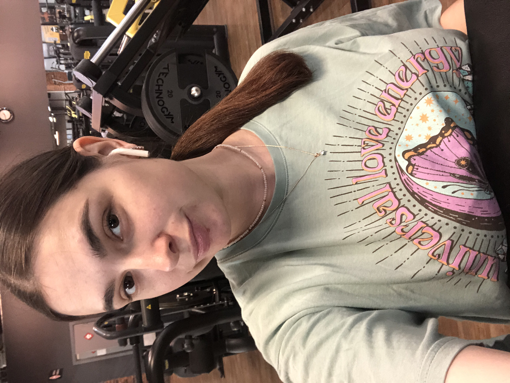 The image shows a person in a gym. The person has brown hair, is wearing a light green t-shirt with a graphic design, and a necklace. The t-shirt graphic includes the words "energy" and "universe". A white wireless earbud is visible in the person's ear. Gym equipment, including weights and a weight machine, is visible in the background. The location is implied to be in a gym in the city of Chaikovsky, Russia.
