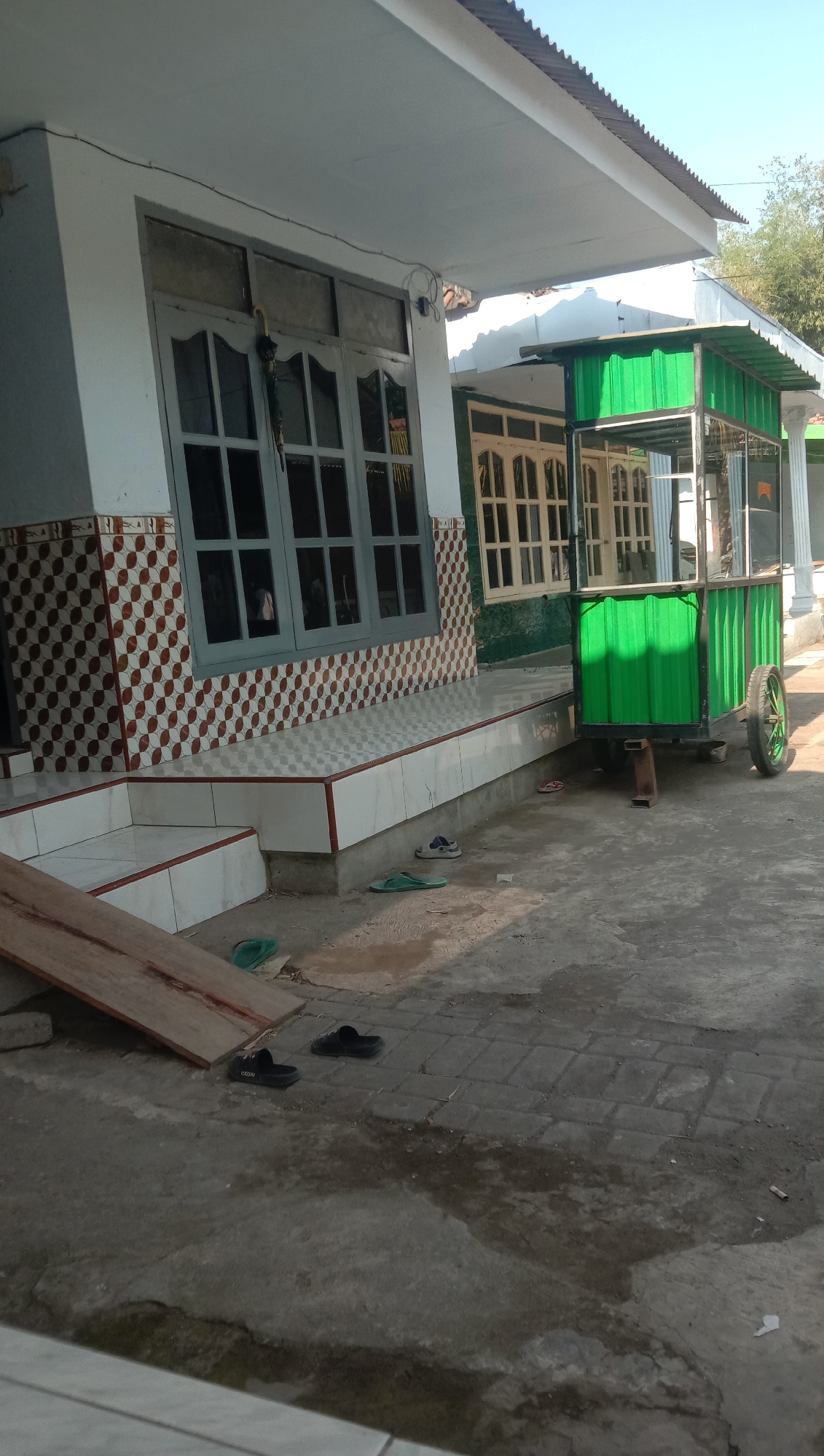 A green cart with a corrugated metal roof is parked on a paved outdoor area in front of a building. The cart has a metal frame and wheels. The building has a white exterior with brown and white patterned tiles on the lower portion of the wall. There are windows with grey frames on the building. Scattered on the ground are a pair of blue and white sneakers, a pair of green flip-flops, and a pair of black sandals. A wooden plank is leaning against a portion of the building's foundation. The scene appears to be in Sidogiri, Indonesia, suggested by the architectural style and the presence of a food cart common in Indonesian street scenes. The lighting indicates it is daytime.