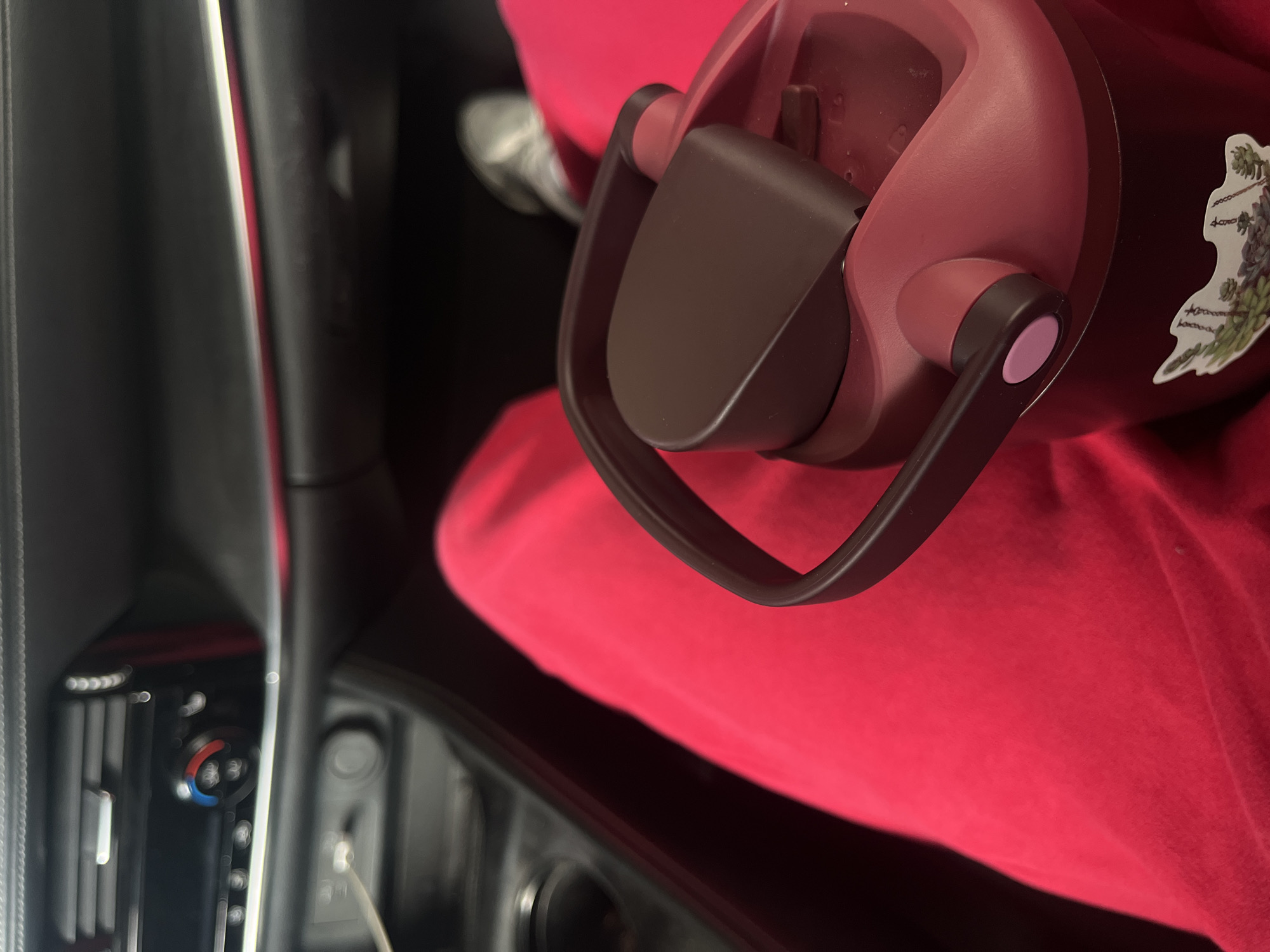 The image shows the interior of a car. A pink water bottle with a brown handle sits on a red surface, possibly a shirt or seat cover. The car's dashboard, including air vents and controls, is visible on the left.