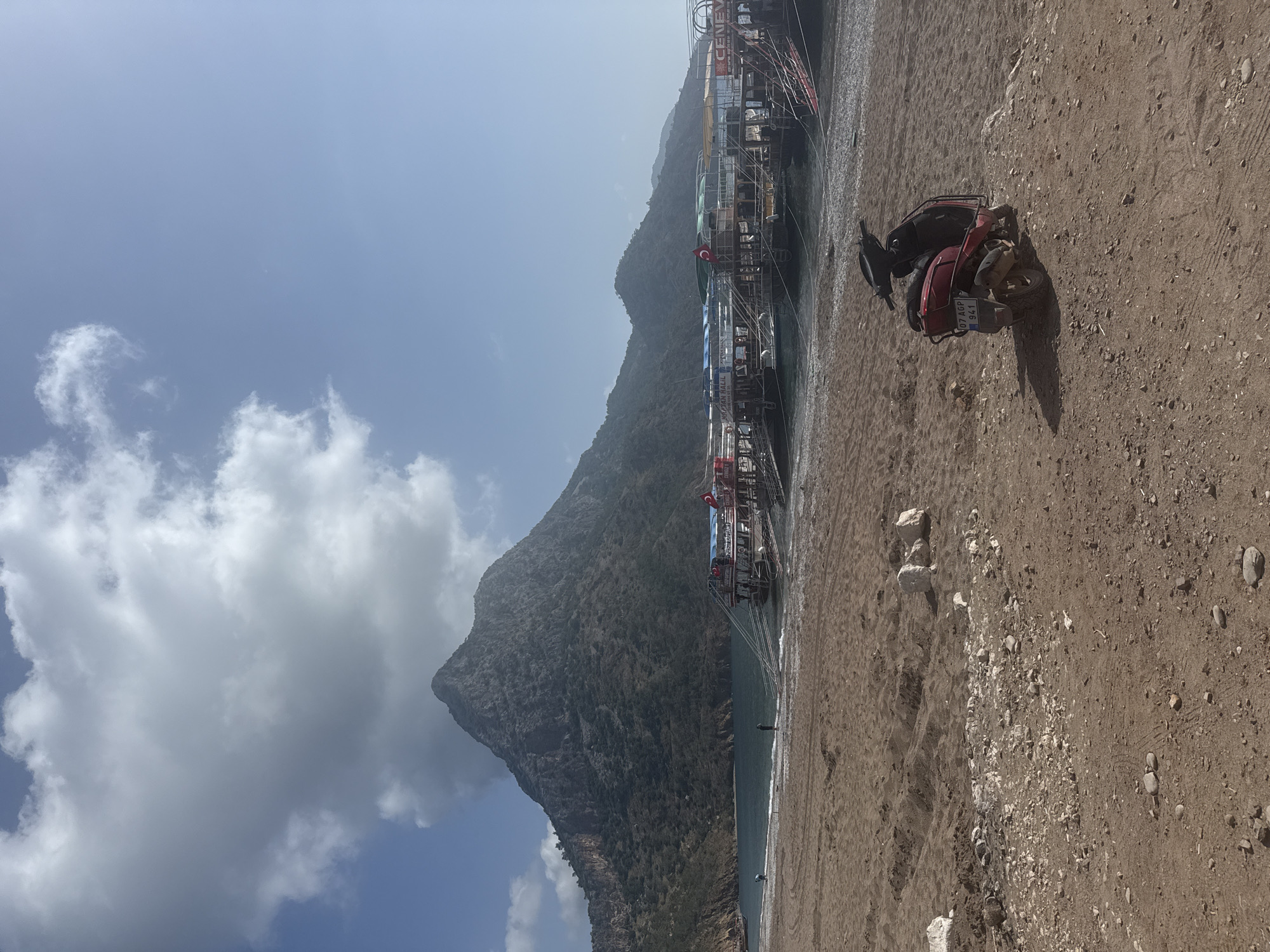 The image shows a view in Kumluca, Turkey, specifically from a beach. A large mountain looms in the background, partly obscured by clouds in the sky. Several tour boats are moored along the shoreline. In the foreground, on the sandy beach, is a parked red four-wheeled ATV. The beach shows signs of vehicle tracks and scattered small rocks. The sky is blue with some white clouds.