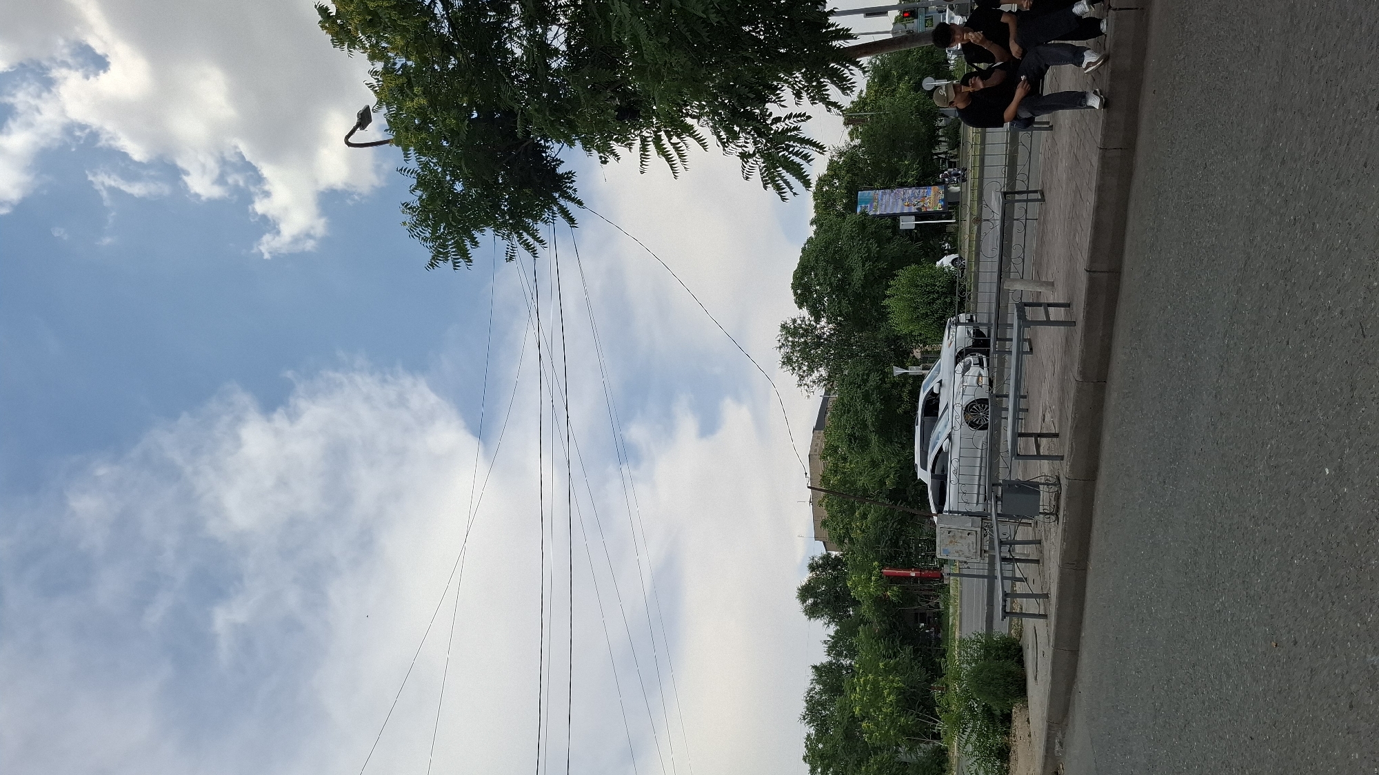 This is a full-frame, eye-level shot of a street scene in Shymkent, Kazakhstan, under a partly cloudy sky. The foreground features an asphalt road leading to a sidewalk with several metal benches and a trash can. A white car is parked on the other side of the sidewalk. Further back, a line of trees creates a green barrier, with a building visible behind the trees. A streetlight stands to the right of the frame, partially obscured by a large tree. Three young men are sitting on one of the benches, eating. One man is wearing a black t-shirt and jeans, another is in a black t-shirt and dark pants, and the third is wearing a black t-shirt, dark pants, and a baseball cap. The sky is a prominent feature, with a mix of white and light blue clouds. Overhead power lines crisscross the sky, adding a sense of urbanity to the scene. The overall impression is a typical day in a moderately populated area of the city.