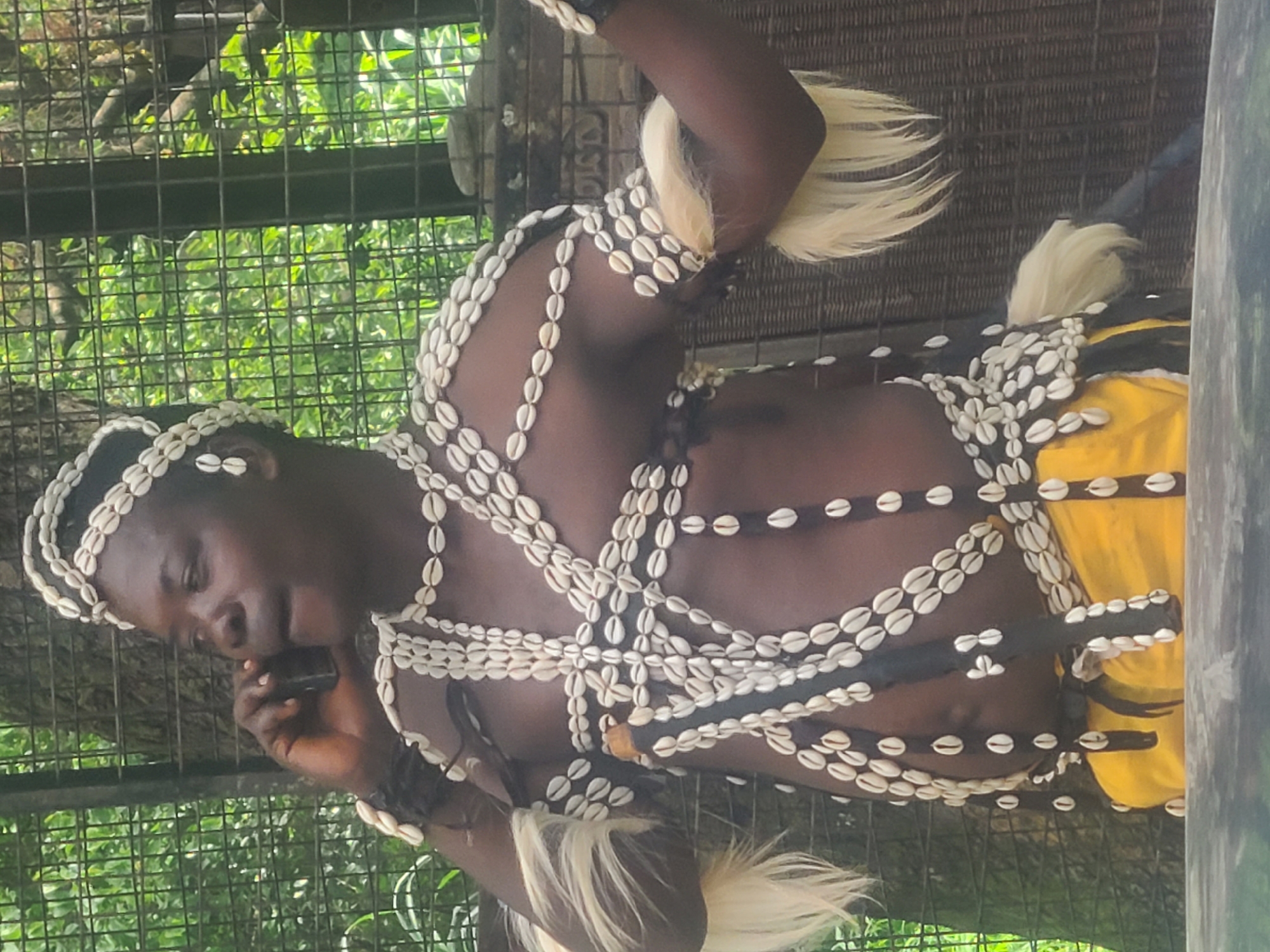 A person wearing traditional attire decorated with seashells and white feathers is standing behind a metal fence, holding a mobile phone to their ear. They are facing to the left, with their mouth slightly open as if speaking. The background consists of lush green foliage and trees, suggesting an outdoor setting, possibly a zoo or a park in Jos, Nigeria. The individual is adorned with multiple necklaces and headwear made of white seashells, and has white tufts of what appear to be fur or hair attached to their arms. A portion of a yellow garment is visible around their waist. The overall lighting suggests it is daytime.