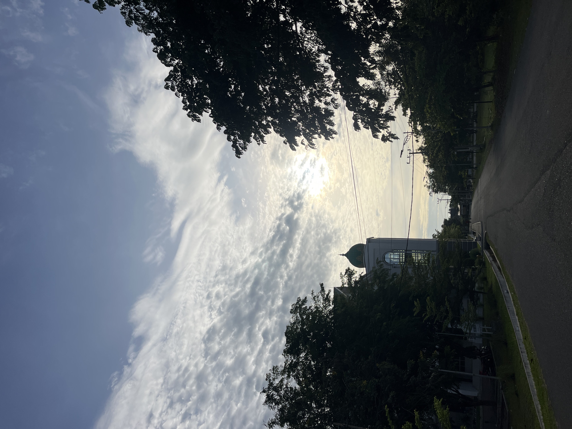A mosque is partially visible behind tree branches on a sunny day. The sky is blue and filled with thin white clouds. A road runs along the right side of the image, lined with trees and utility poles. The location is presumed to be in Lhokseumawe, Indonesia.