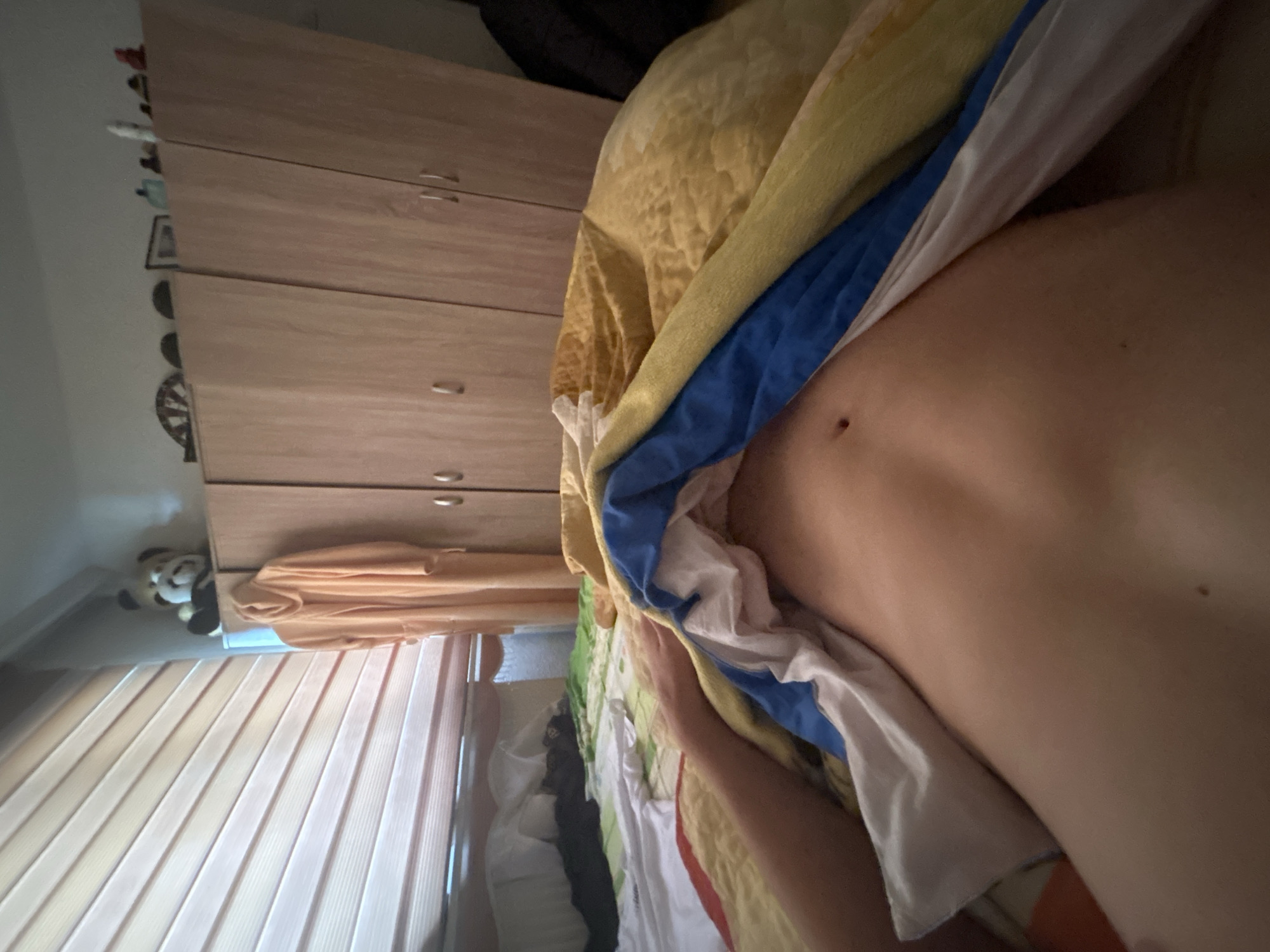 The image shows a close-up view of a person lying in bed. The person is partially covered by a yellow, blue, and white blanket. The blinds on the window are visible, and there's a wardrobe with clothes in the background. A toy is visible on the side. The location is likely a bedroom. The location is assumed to be within Zlatograd, Bulgaria.