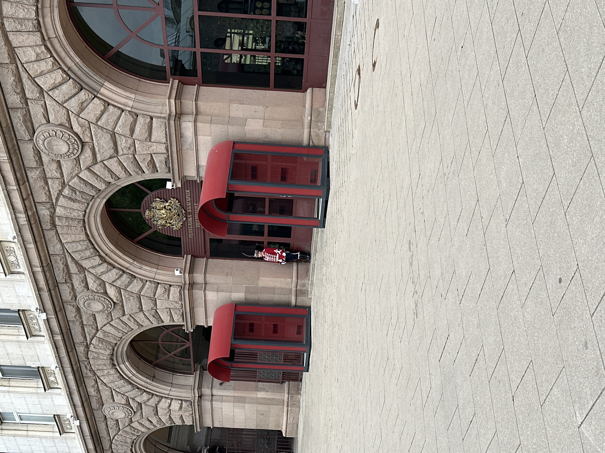 The image shows the entrance to the Presidency building in Sofia, Bulgaria. The building's façade is made of light-colored stone and features arched windows and decorative elements. A guard in traditional Bulgarian attire is standing at the entrance, positioned between two red awnings. The sidewalk in front of the building is paved with light-colored stone tiles. The photograph is taken from a ground-level perspective and rotated.