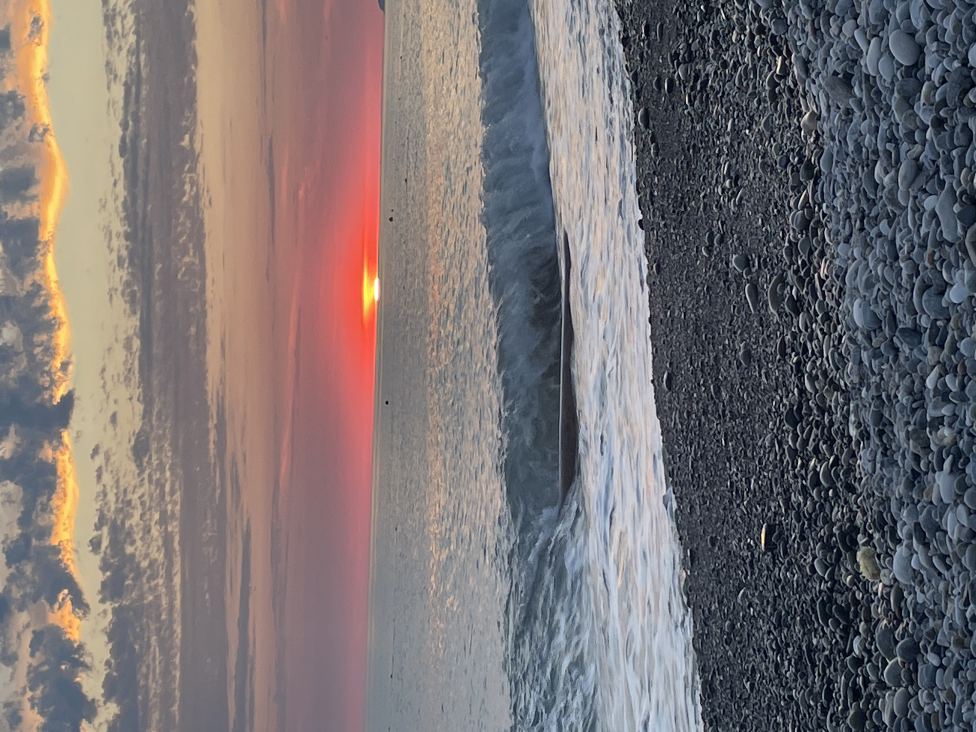 A wide shot captures a tranquil beach scene at sunset, with the sun appearing as a bright orange orb just above the horizon line of the calm ocean. The sky is a gradient of colors, transitioning from a deep pinkish-red near the sun to lighter pinks and purples, with scattered clouds tinged with orange and gray. Gentle waves are breaking on the shore, creating white foam that washes onto the pebbled beach. The foreground is dominated by the textured surface of the beach, covered in a variety of smooth, gray and dark-colored pebbles. The overall atmosphere is peaceful and serene, with no people or animals visible in the frame.