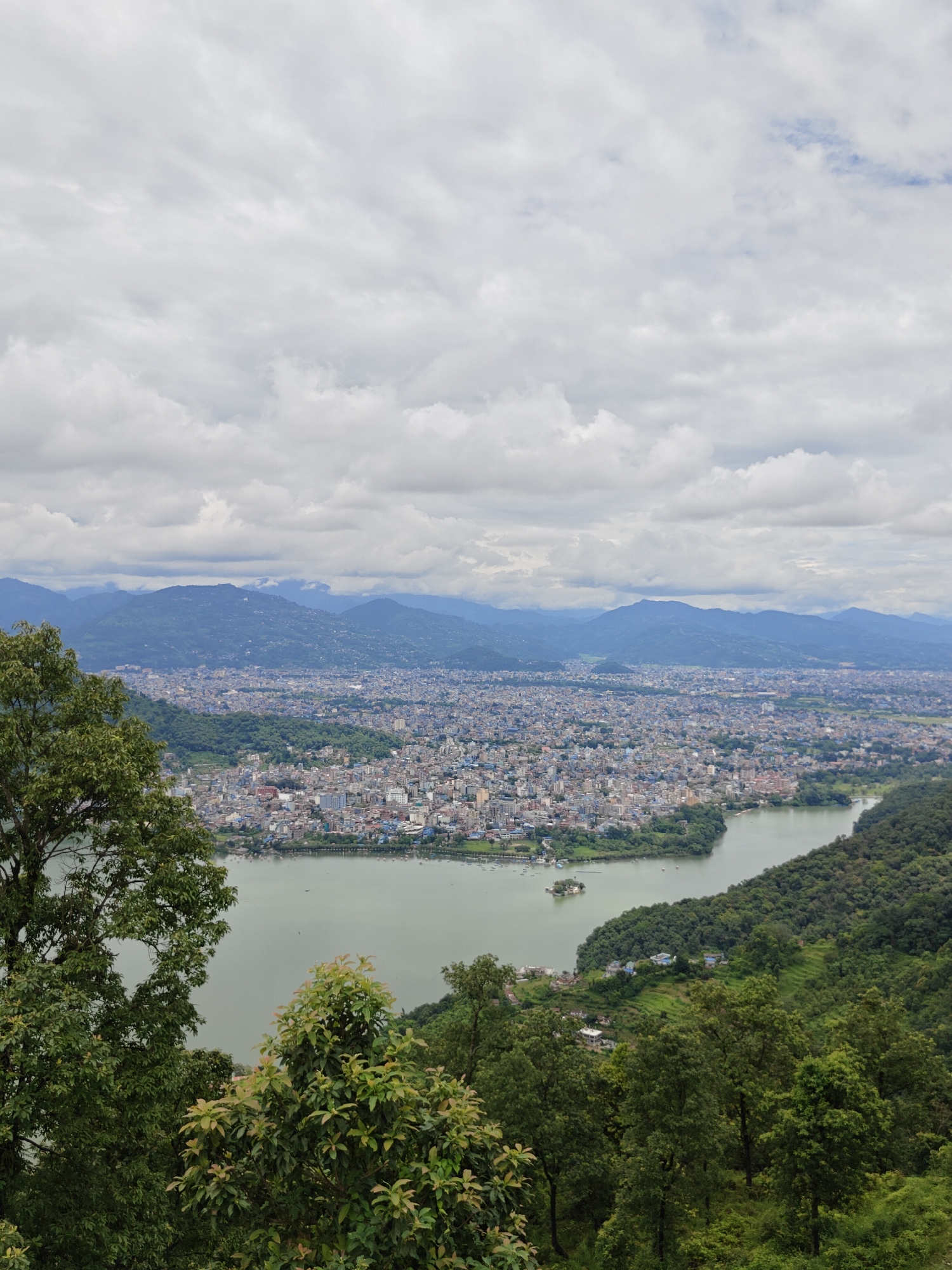 The image captures a panoramic view of Pokhara, Nepal, featuring Phewa Lake and the surrounding cityscape. In the foreground, lush green trees and foliage create a natural frame, with a large tree on the left and dense vegetation on the right. The middle ground is dominated by the tranquil waters of Phewa Lake, a narrow strip of land, and a small island with a pagoda. Beyond the lake, the city of Pokhara sprawls across the landscape, characterized by a dense collection of buildings with varied architectural styles and colors, predominantly in shades of white, grey, and blue. The city extends towards the horizon, where rolling hills and mountains are visible, shrouded in a misty haze under a cloudy sky. The clouds are predominantly grey and white, suggesting an overcast day. There are no discernible people or animals in the image. The overall scene depicts a serene blend of natural beauty and urban development, offering a high-angle perspective of the city.