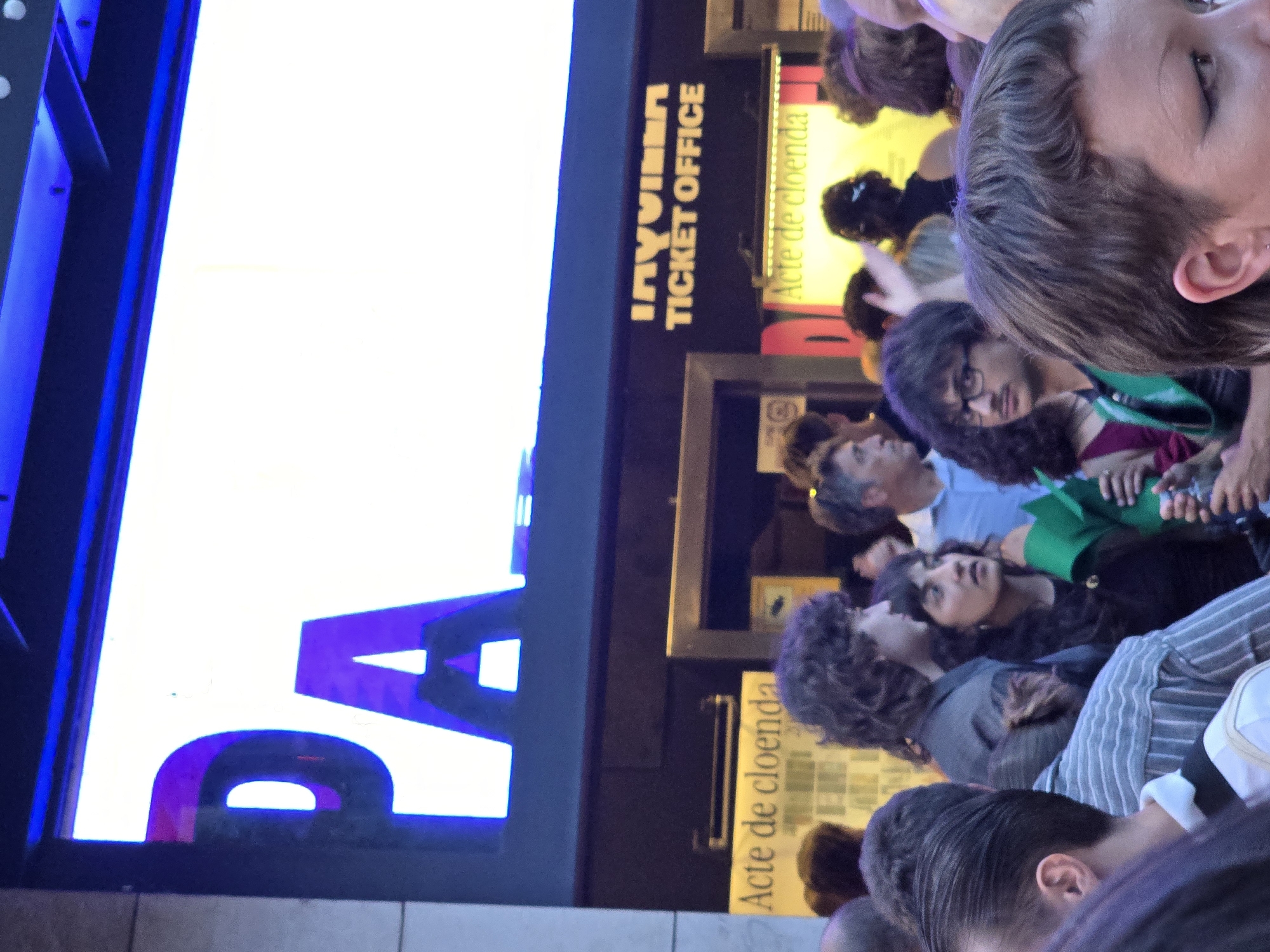 This media file depicts a crowd of people gathered in what appears to be an outdoor or semi-outdoor setting, likely at night given the artificial lighting. The scene is characterized by a mix of individuals, some looking up and others looking down or at the camera.
The primary focus is on the people in the foreground and mid-ground, with a strong emphasis on their faces and hairstyles. The individuals have diverse hair colors and textures, with some exhibiting curly hair and others straighter styles. Their expressions are varied, ranging from attentive to contemplative.
Visible in the background are signs and signage that provide clues about the location. One sign reads "TICKET OFFICE" in large, blocky letters, suggesting a venue that sells tickets, possibly for an event or attraction. Another sign, written vertically, reads "Acte de cloenda" which is Catalan for "Closing Act" or "Closing Event." This suggests the gathering might be for the conclusion of something.
The presence of bright, possibly neon, blue lighting on the left side of the image, and the glowing yellow and red hues from the signs on the right, create a vibrant and somewhat chaotic atmosphere. The overall impression is one of a bustling public space, perhaps a street festival, a concert venue, or a cultural event in Barcelona, Spain. The time of day is likely evening or night due to the illumination. The emotions conveyed by the people are a mixture of anticipation and engagement with whatever is happening around them.