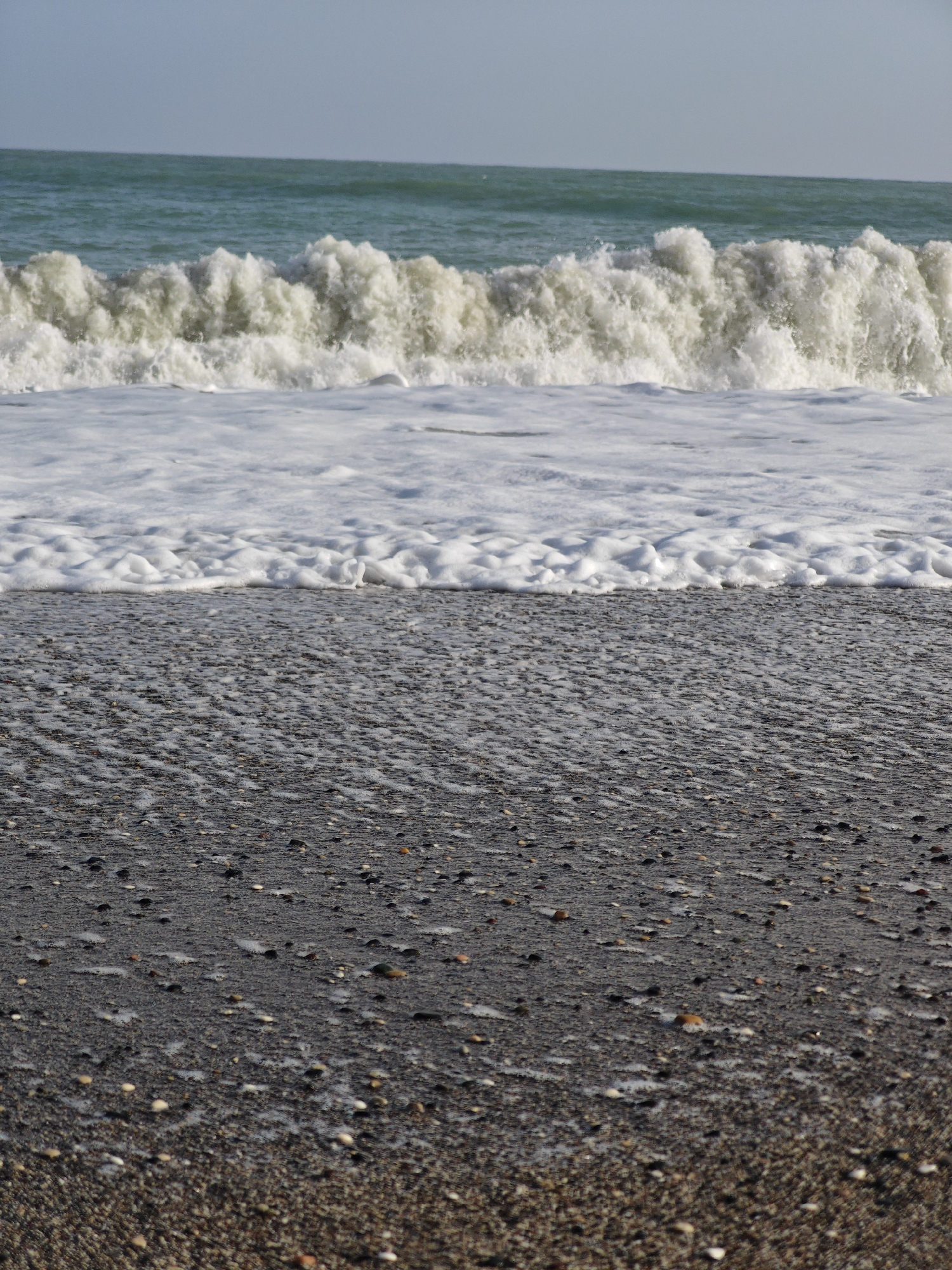 A close-up shot captures the coastline near Demre, Turkey. The foreground features a dark, pebbled beach, transitioning to a stretch of wet sand reflecting the retreating waves. A layer of foamy surf marks the boundary between the beach and the turquoise sea, where a large wave is breaking, its crest a mass of white foam. The horizon is visible as a faint line under a light blue sky. No people or other objects are visible in the scene. The overall impression is one of the raw power and beauty of the ocean meeting the shore.