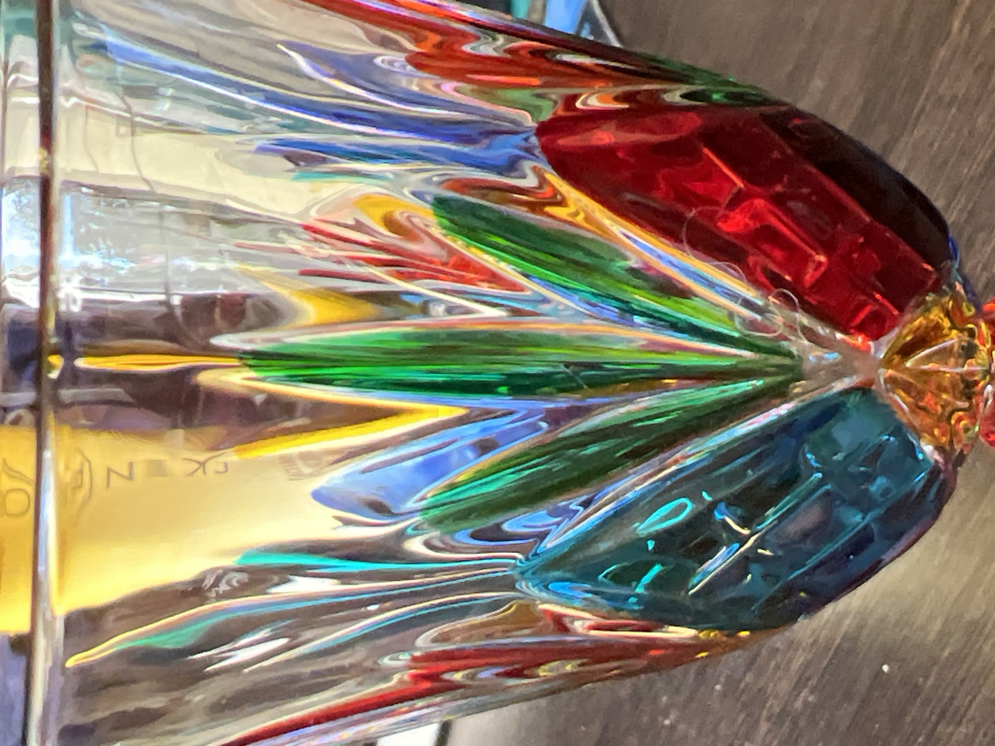A close-up, eye-level shot shows the intricate details of a multi-colored, cut-glass vase. The vase is positioned on a dark wood surface, with the background blurred and out of focus, displaying a spectrum of iridescent colors. The vase itself is faceted, with a pattern of pointed, petal-like shapes in vibrant hues of red, blue, green, and yellow. The light refracts through the glass, creating a dazzling display of color and reflection. The overall impression is one of richness and artistry. The image does not contain any elements that would allow for the identification of the city of Sinyavino, Russia.