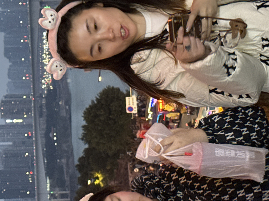 This image captures a nighttime or dusk urban scene in Yuzhong District, Chongqing, China, observed from an elevated viewpoint.
In the foreground, two individuals are partially visible. The central figure on the right, an East Asian female, wears a pink headband adorned with two fuzzy white animal ears. Her mouth is open as if speaking or reacting. She is dressed in a light-colored, possibly cream-colored, puffy jacket featuring a repeating dark "NY" pattern. Her left hand holds a dark, rectangular object, possibly a mobile phone, from which a small, brown, animal-shaped charm dangles by a strap. A tattoo is visible on her left hand near the index finger. The individual on the far left, also an East Asian female, is mostly obscured, but her hands are visible holding two clear plastic bags. Each bag contains a beverage, and one displays the Chinese characters "蜜雪冰城" (Mixue Bingcheng) with a red straw visible. This individual wears a dark jacket with a repeating white "NY" pattern.
The background reveals a sprawling cityscape, dominated by numerous high-rise buildings with illuminated windows under a dark or overcast sky. A wide river, likely the Jialing or Yangtze River, flows through the city, spanned by a large bridge with multiple lanes. Vehicle lights, indicating active traffic, are visible across the bridge. Below the foreground subjects, at street level, a dense crowd of people is visible alongside several brightly lit vehicles, including what appear to be commercial or tour buses. A circular blue street sign with a white horizontal bar is also present at this lower level, and dark green trees and foliage line the riverbank in the mid-ground. Artificial lighting from streetlights, buildings, and vehicles illuminates the entire scene.