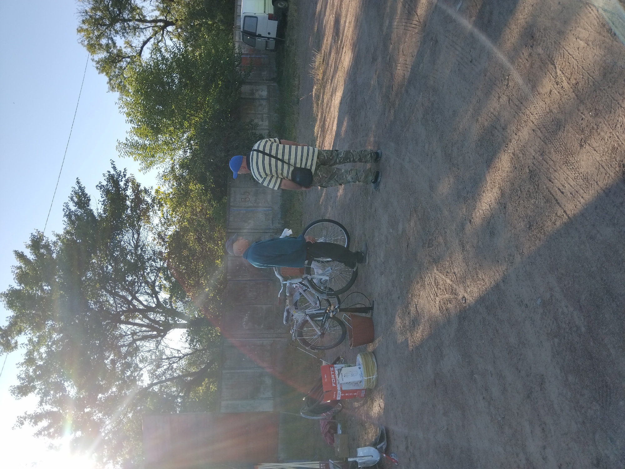 Two men are standing in an outdoor lot next to a concrete fence. The man on the left is wearing a blue jacket and a cap, and is looking to the right. The man on the right is wearing a yellow and black striped shirt, camouflage pants, and a blue cap, and is looking to the left. In front of the man on the left are two bicycles, one is partially disassembled. To the left of the bicycles are boxes and coils of yellow cable. To the right of the bicycles is a bucket and a bicycle pump. In the background, a white van is parked next to the fence. The ground is dirt and gravel. The scene is illuminated by bright sunlight. The location is the city of Irshansk, Ukraine.