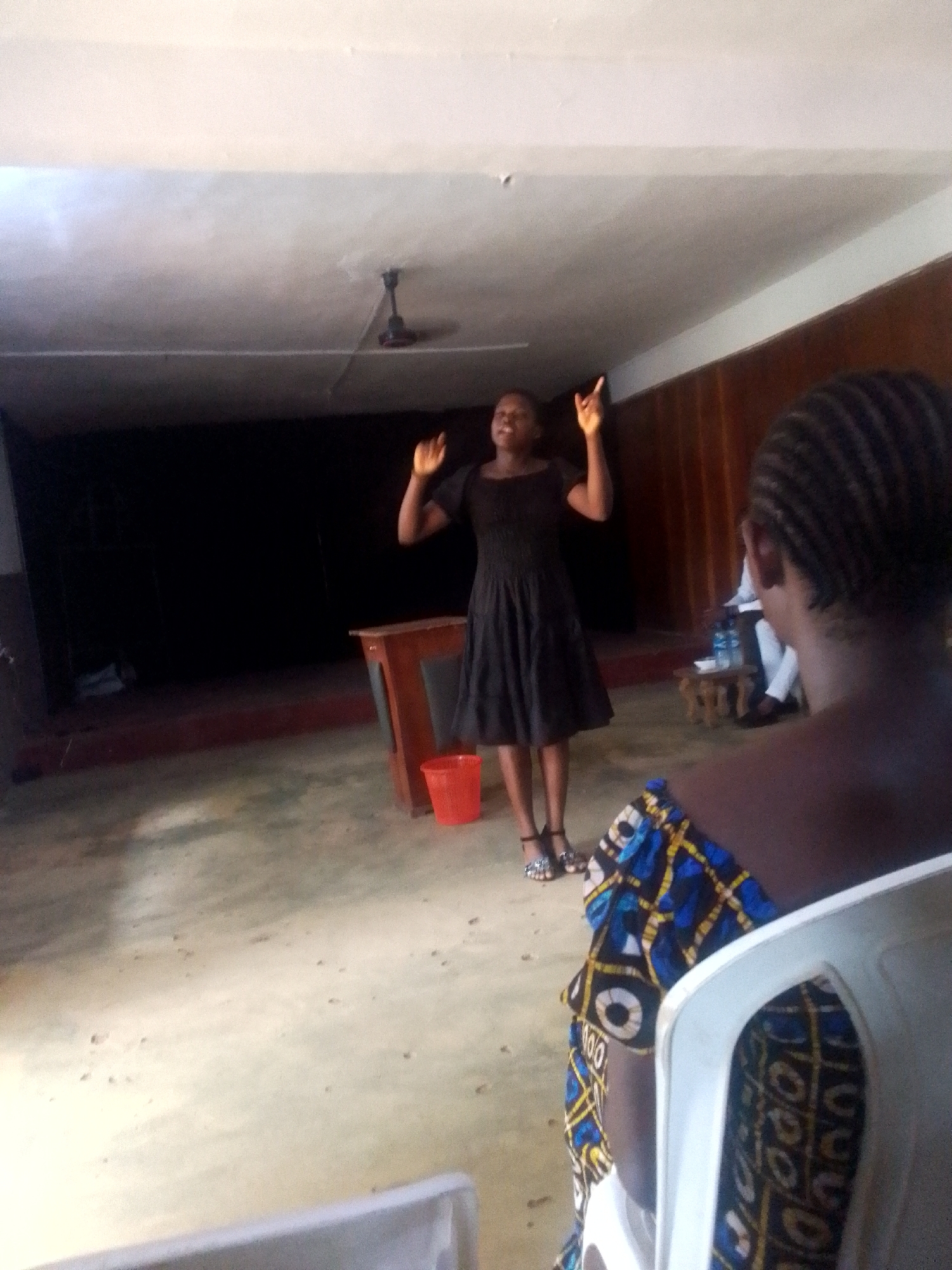 The photo depicts a woman wearing a black dress and sandals standing on a stage-like platform in an indoor setting. She has her arms raised, with her fingers pointed upward. Behind her is a small wooden table and a red plastic bucket. A dark backdrop is visible behind the table. In the foreground, a person is seated in a white plastic chair, facing away from the camera. The person is wearing a colorful patterned top. Other people are partially visible in the background. The setting appears to be a simple hall, possibly a community center or church hall. A ceiling fan is visible on the ceiling. The location is Unknown, Unknown.