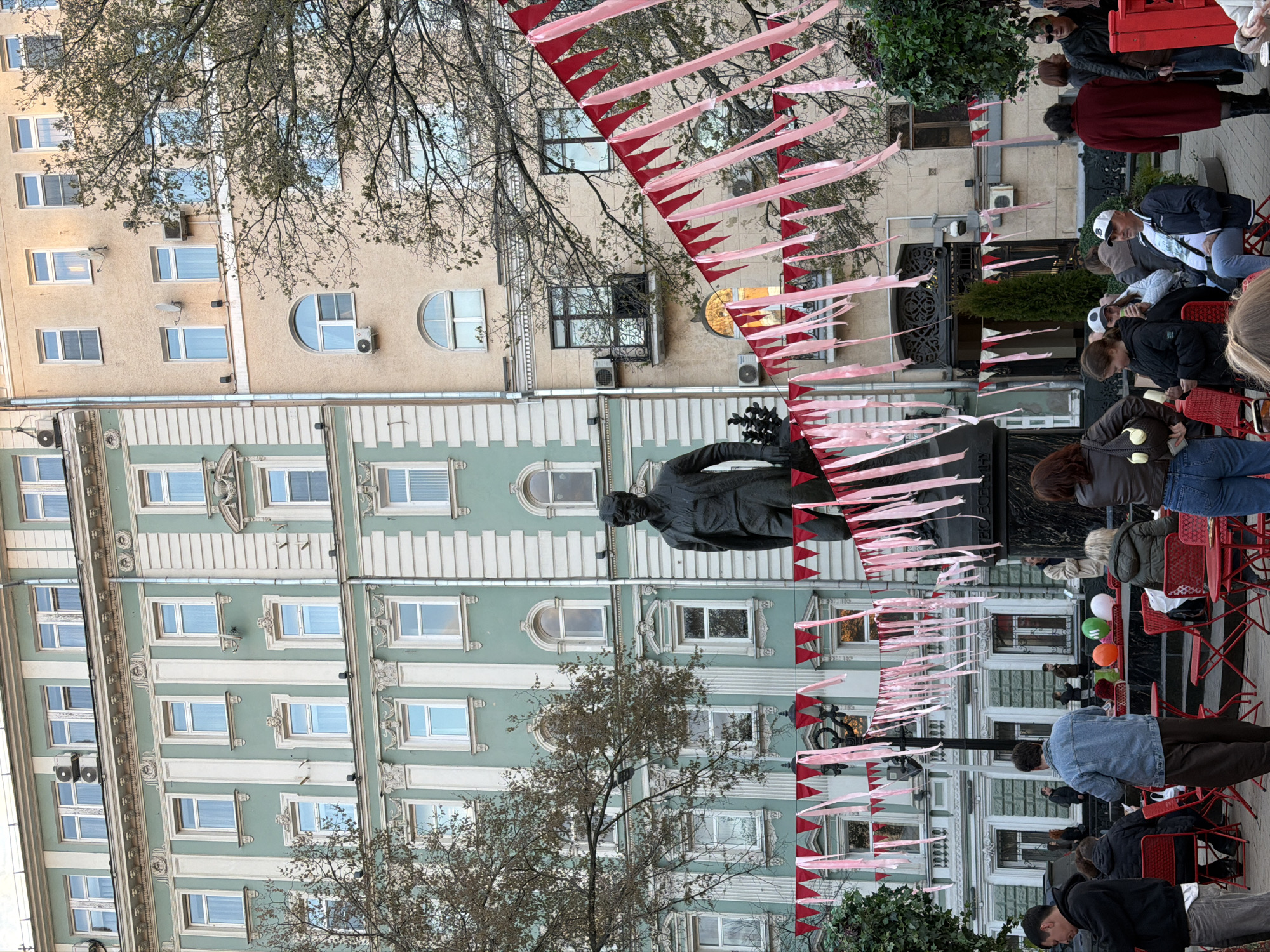 The image depicts a street scene in Moscow, Russia, featuring a statue in front of a multi-story building with a light green and white facade. There are rows of windows on each floor and ornamental designs above the windows. A second building with a beige facade is visible on the right. Red and pink pennant banners stretch across the street, above the statue. People are seated on red chairs in front of the building, appearing to be watching something. There are also people standing and walking around. A tree with bare branches is visible in the foreground.