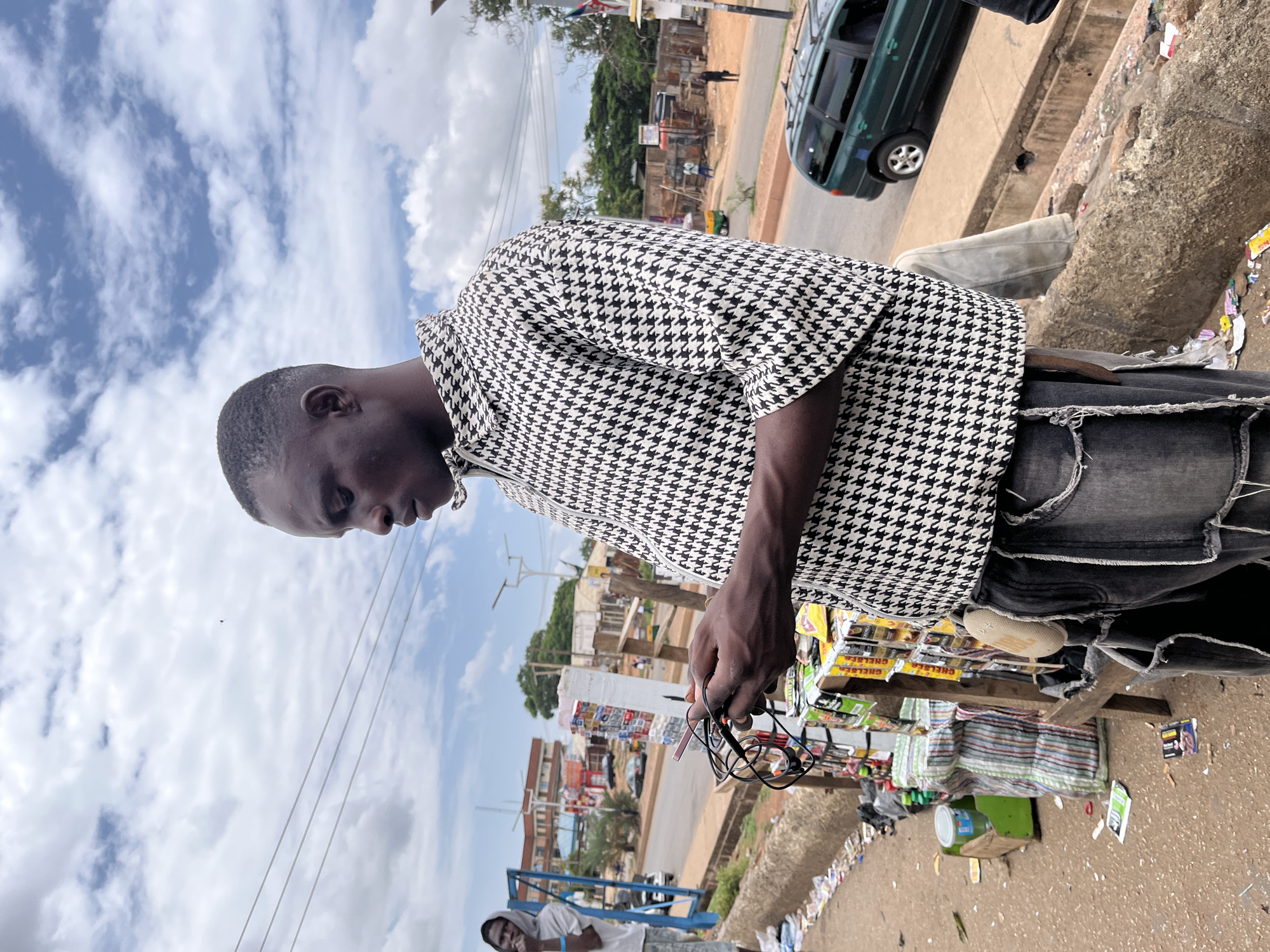 The image depicts an outdoor street scene in Jos, Nigeria, characterized by urban infrastructure and a makeshift vendor stall under a partially cloudy sky. The primary subject is a dark-skinned male standing in the foreground, facing slightly left with his gaze directed downwards. He wears a black and white houndstooth pattern long-sleeved shirt with a zippered collar and distressed dark grey jeans from which a light-colored, textured pouch hangs from the right hip. His hands are occupied with a tangled pair of black wired earphones or earbuds, which he appears to be examining.
To the subject's left, a rudimentary wooden stall is present, displaying various packaged consumer goods such as snacks, candies, and possibly beverages, including items labeled "Chelsea." Several striped sacks are stacked on the ground near the stall. The ground in the immediate vicinity is unpaved, consisting of dirt and gravel, and is heavily littered with wrappers, papers, and plastic debris. Further left, a second dark-skinned male, partially visible, stands near a blue metal frame, wearing a light grey hooded shirt and shorts.
Beyond the foreground, a paved road extends into the distance, bordered by concrete drainage channels. A dark green station wagon-type vehicle is visible on the road to the right. Various buildings with varying states of completion and reddish roofs line the street in the midground and background. Multiple power lines traverse the upper portion of the sky, which exhibits a mix of white clouds and blue patches. A utility pole with a small flag, possibly the Nigerian flag (green and white), is visible on the right side of the road. Distant figures are discernible on the far side of the street. The overall environment suggests a bustling, somewhat unkempt urban setting.