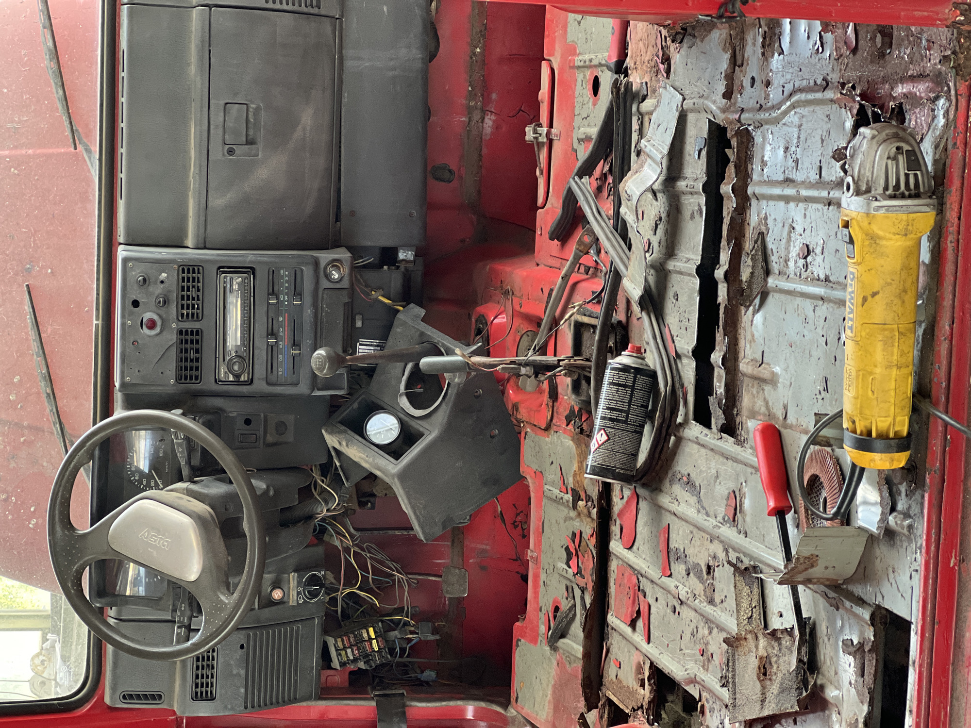 The image shows the interior of a red vehicle, likely a Suzuki Samurai, in a state of disrepair and undergoing restoration. The floorboards are heavily rusted, with pieces of metal and red paint flaking off. Tools, including an angle grinder and a can of spray paint, are scattered on the floor. The dashboard, steering wheel, and center console are visible, but appear dusty and worn. Wires are exposed and hanging from various parts of the interior. The scene suggests a DIY restoration project, likely taking place in a workshop or garage. The location in Lanžhot, Czechia is not evident from the image itself but has been provided as context.