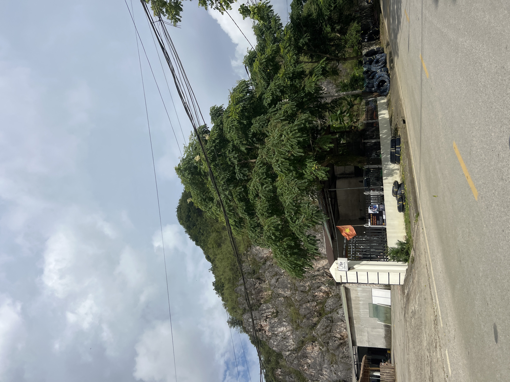 The image shows a scene in Quản Bạ Commune, Vietnam, characterized by a steep, rocky mountain face covered in vegetation. In the foreground, a paved road runs horizontally, with yellow dividing lines. To the left of the road, there is a building with a concrete wall and a section that appears to be a workshop or storage area, with some glass panes leaning against the wall. Adjacent to this building is a white pillar with black stripes, bearing a sign that reads "Hạn Coffee & Tea." A black fence encloses a small area behind the pillar, where a Vietnamese flag is flying. Several large trees with lush green foliage are prominent in the middle ground, obscuring parts of the mountain. To the right of the fence, a pile of black coiled pipes or hoses is stacked on the ground, and more are visible further along the roadside. Overhead, a network of power lines crisscrosses the sky, which is partly cloudy. The overall environment suggests a semi-rural or commercial area bordering a natural landscape. No people are visible in the image.