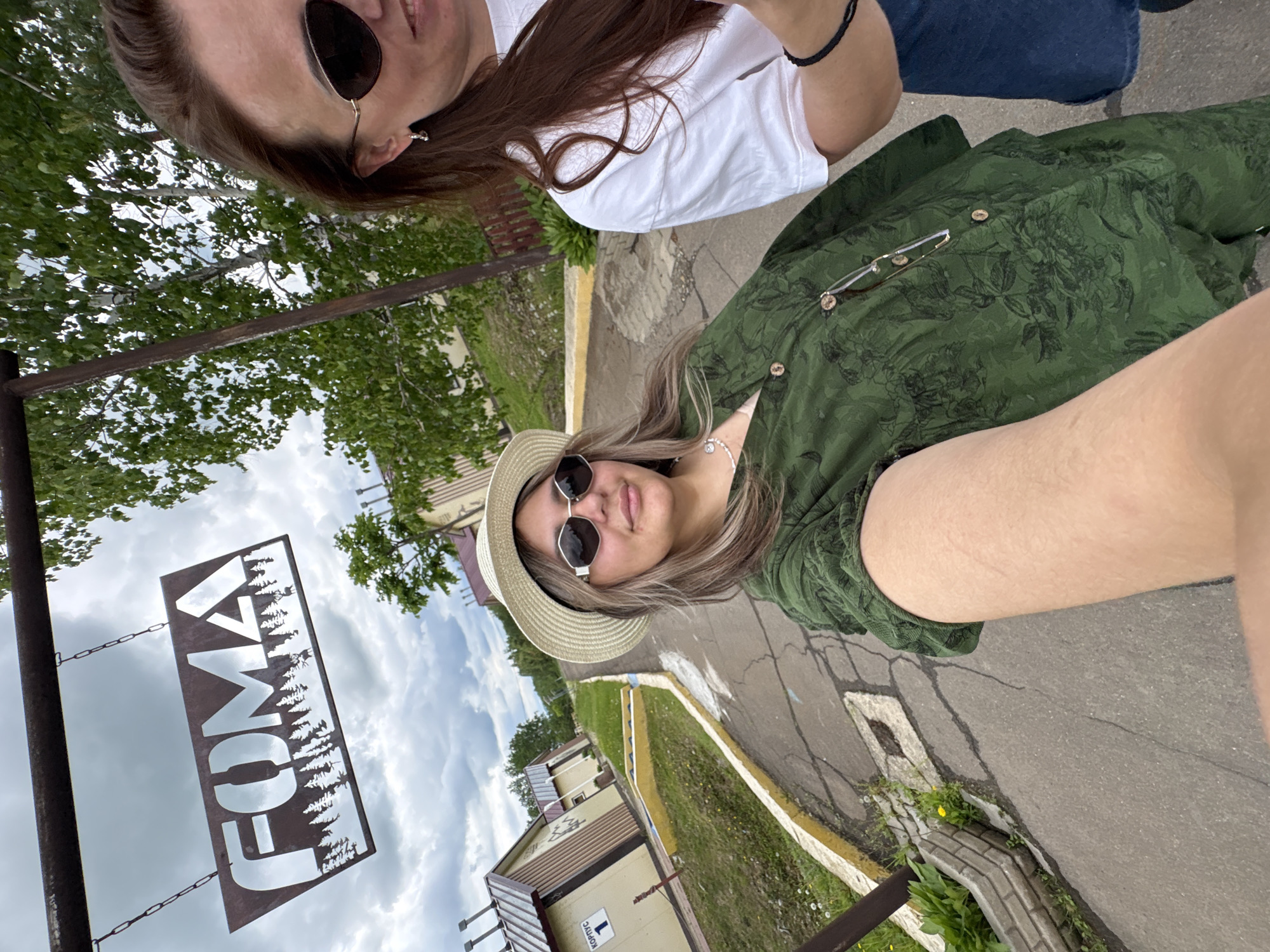Two women are standing on a paved path. The woman on the left is wearing a green patterned dress, a straw hat, and sunglasses. The woman on the right is wearing a white t-shirt and sunglasses. A sign reading "FOMA" hangs above them. In the background, there are several small buildings and trees. The location is a tourist complex in the city of Birobidzhan, Russia. The sky is cloudy.