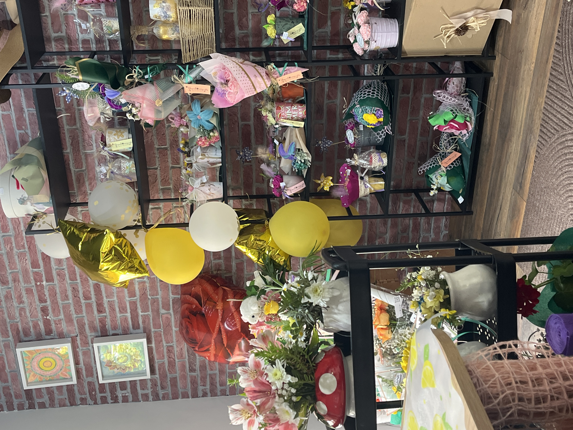 The image shows the interior of what appears to be a gift shop, likely in Burgas, Bulgaria given the prompt context. The scene is vertically oriented, capturing a retail display with a focus on floral arrangements and gift items.
A black metal shelving unit displays various packaged bouquets, arrangements in containers, and decorative items. The top shelves feature many wrapped bouquets, some with ribbons, flowers, and tags. Beneath, several white and yellow balloons are suspended from the shelves, along with a gold, star-shaped balloon and a red balloon shaped like a rose.
The lower shelves contain fresh floral arrangements in vases, as well as other shop items, including decorative mushrooms. In the bottom left corner, two framed pictures adorn a section of the wall, which is composed of red bricks. The overall lighting is ambient. The style and presentation of the displayed items suggest the location to be a gift shop specializing in floral gifts.