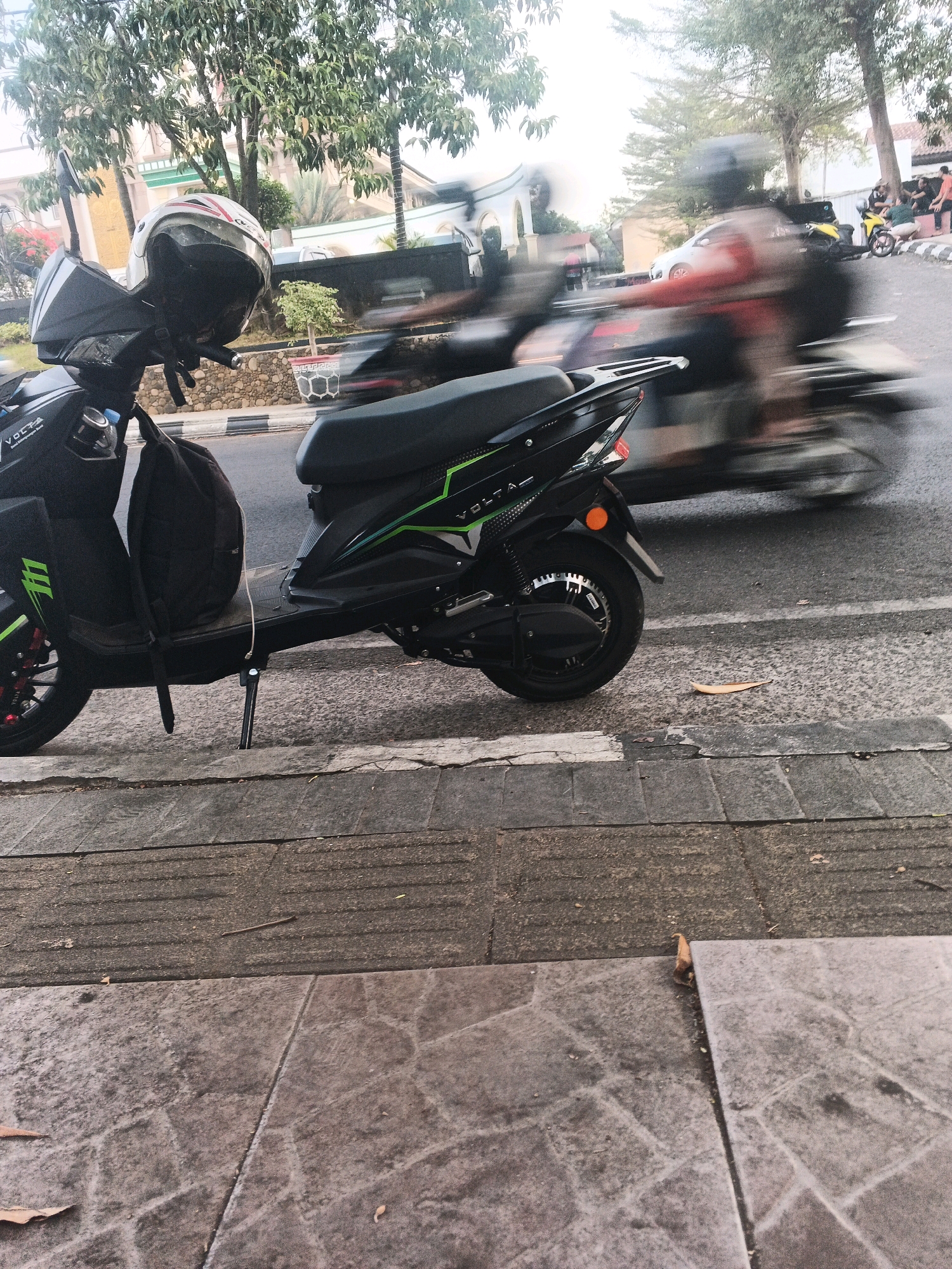 In Sidomulyo, Indonesia, a black electric scooter is parked on a sidewalk next to a street. The scooter has a black backpack strapped to its seat. A white and red helmet rests on the handlebars. In the background, traffic is moving, with several motorcycles and a car blurred due to motion. Trees and buildings line the street. The sidewalk is made of gray paving stones with textured strips.