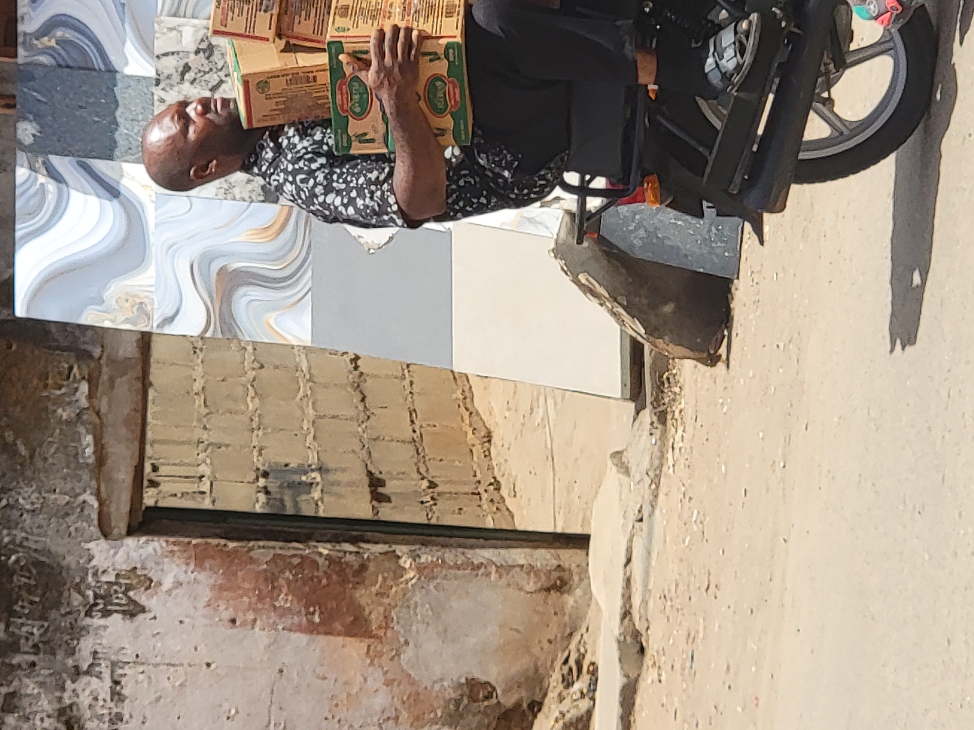 This outdoor photograph, taken in Maiburiji, Nigeria, captures a man transporting goods on a motorcycle under bright daylight. The central figure is a dark-skinned man, bald and wearing a black and white patterned long-sleeved shirt and dark trousers. He is seated on a black motorcycle and is holding a stack of several light brown cardboard boxes in front of him. Each box features a prominent green circular logo with the word "GLORY" written in white, alongside other red and black text that is illegible. The man is looking slightly upwards with a neutral expression.
The setting behind him features contrasting architectural elements. To the immediate left of the man, a wall is covered with large, polished tiles in shades of white, grey, and blue, adorned with swirling patterns. Further left, an older section of a building displays rough, weathered brick or concrete walls with visible wear and tear, and a dark rectangular opening, possibly a doorway or window. The ground is a dusty, light-colored surface, and strong shadows cast by the motorcycle indicate a sunny day. The scene depicts a moment of everyday activity, likely related to delivery or trade, in what appears to be a semi-urban or commercial area.