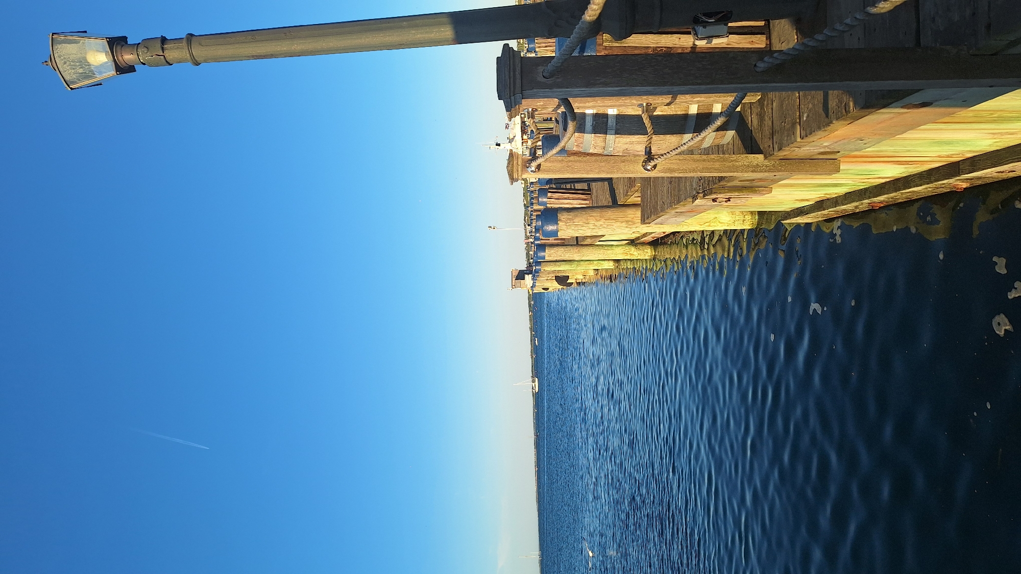 A vertical, eye-level shot captures a wooden dock extending into a calm, blue body of water under a clear blue sky. A single jet stream is visible in the sky. On the right side of the frame, a tall, dark lamppost with a hexagonal glass shade stands at the edge of the dock. The wooden planks of the dock show signs of wear and weathering, with some displaying greenish discoloration. Rope railings line the edge of the dock, secured by thick wooden posts. In the distance, several sailboats and other vessels are scattered across the water, and a small building with a peaked roof is visible on the far side of the water. The overall scene suggests a serene waterfront setting, likely a harbor or bay.