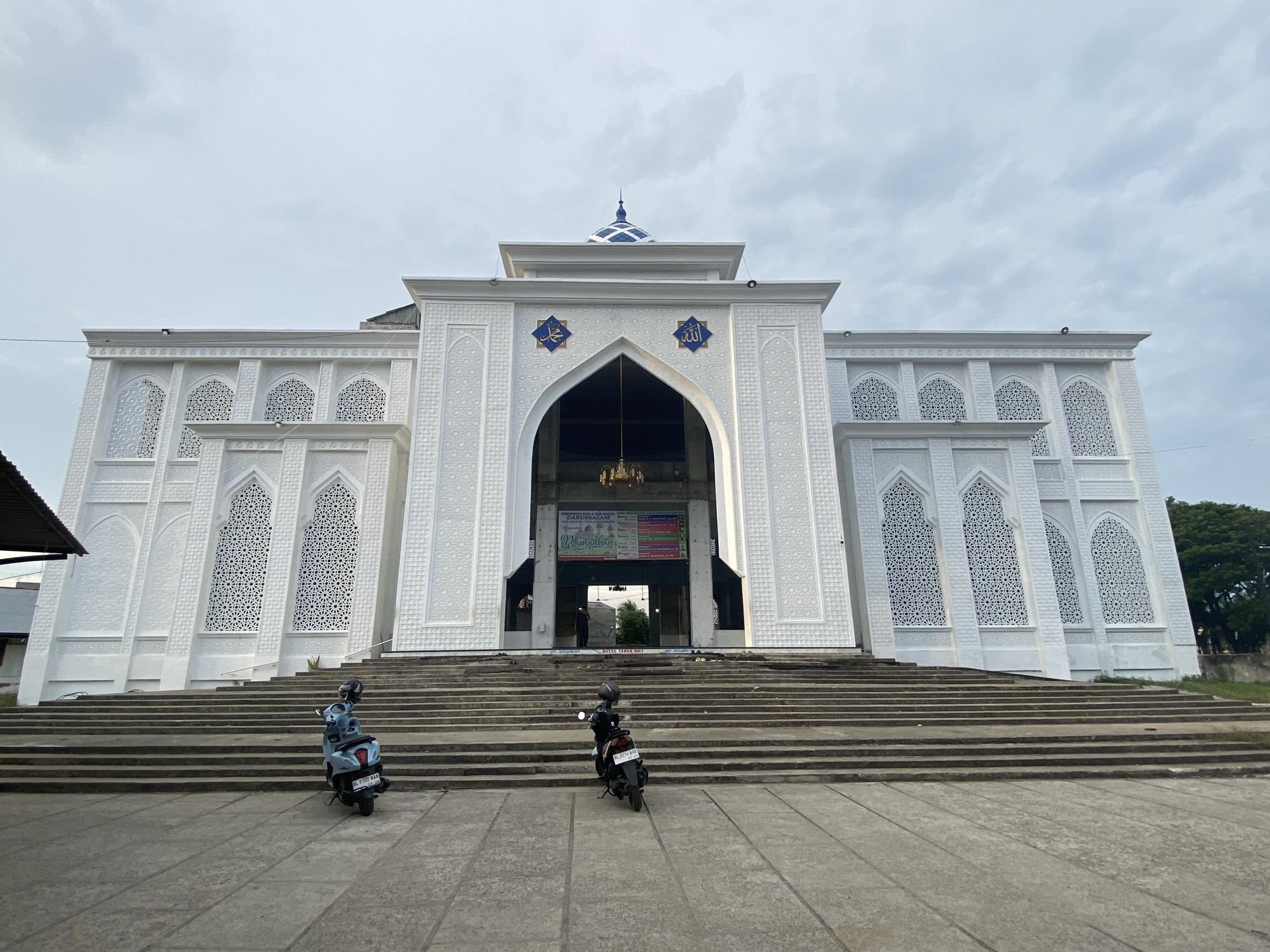 The image shows the facade of a white mosque with intricate geometric patterns on its exterior walls and arched windows. The mosque has a central arched entrance leading into the building, with a blue dome topped by a spire on the roof. Two motorcycles are parked on the steps leading up to the mosque. One is a light blue scooter on the left, and the other is a black motorcycle on the right. The sky above is overcast with clouds. The scene is set in Lhokseumawe, Indonesia, as indicated by the license plates on the motorcycles.