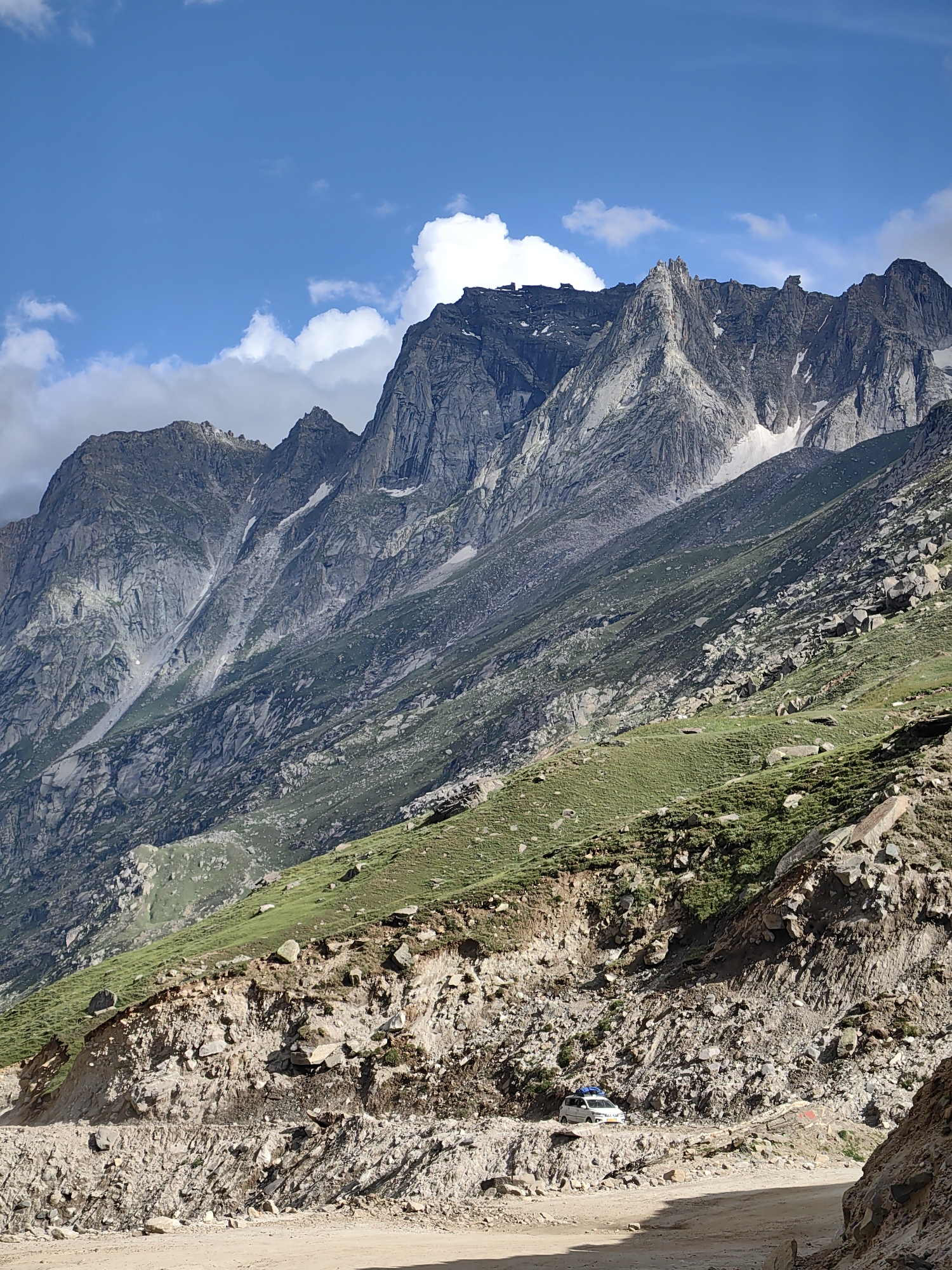 The image depicts a mountainous landscape in the vicinity of Bashisht, India. The foreground features a dirt road carved into a rocky hillside, with a small white car parked on the side. The midground is dominated by a steep, green, grassy slope dotted with rocks and boulders. Beyond this, a majestic mountain range rises, characterized by rugged, gray rock faces, patches of snow, and dramatic peaks under a bright blue sky with scattered white clouds. The scene suggests a remote, high-altitude environment.