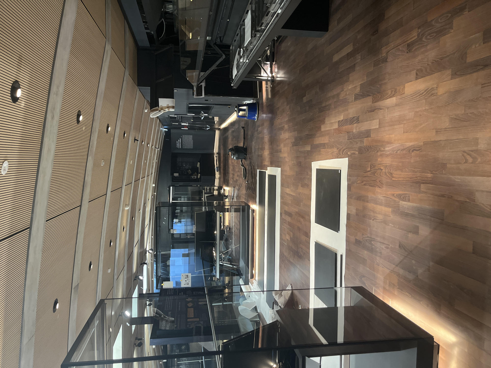 The image depicts the interior of a museum exhibition space in Cologny, Switzerland. The room features a polished wooden floor and a ceiling with a slatted wood finish. Various display cases made of glass and dark metal are present throughout the space, some containing exhibits or equipment. In the mid-ground, a vacuum cleaner and a small blue stool are visible on the floor, suggesting ongoing cleaning or setup activities. The lighting appears to be a mix of overhead fixtures and accent lighting along the display cases. The overall impression is one of a modern, well-lit exhibition area undergoing preparation or maintenance.