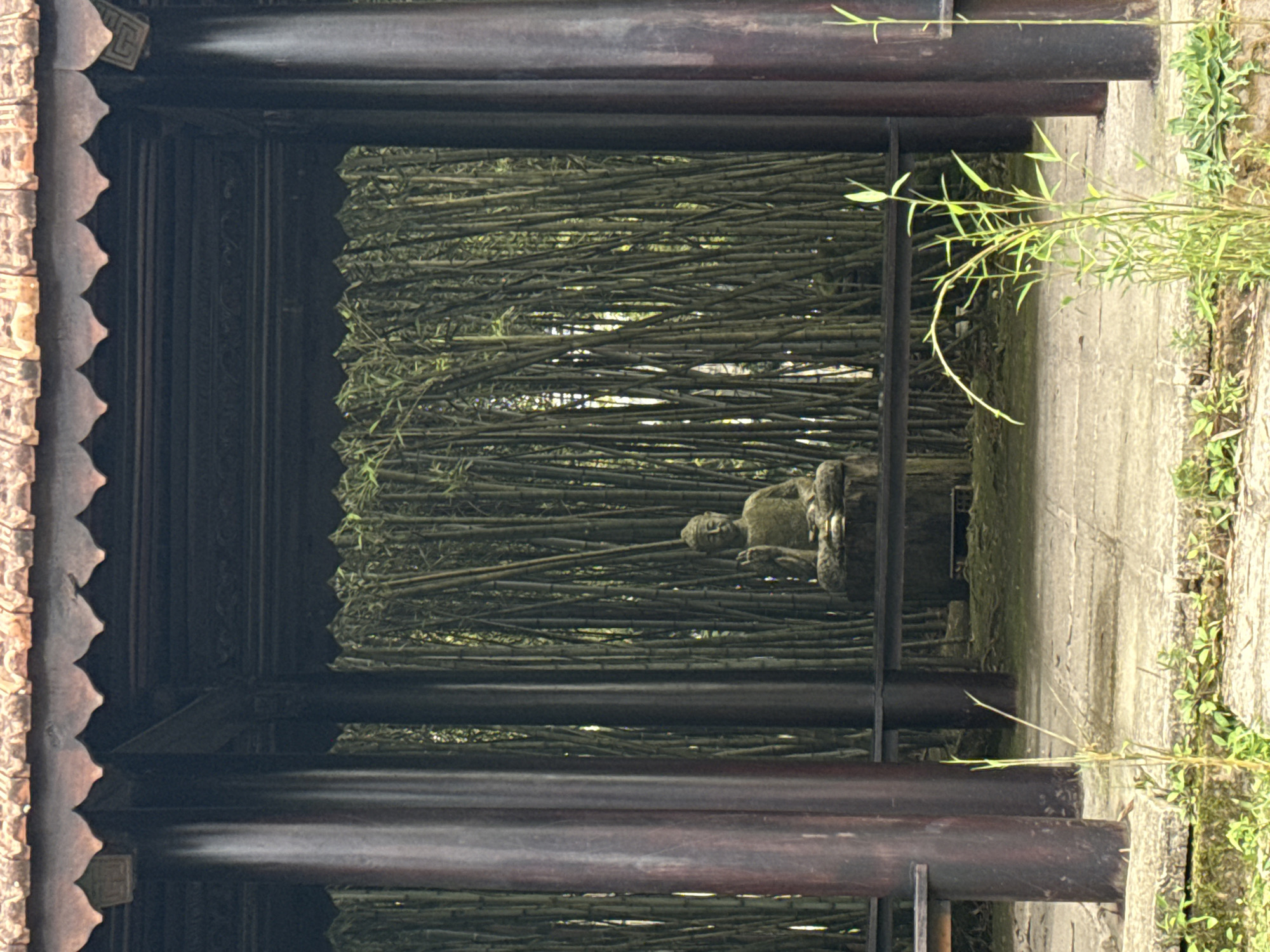 A serene outdoor scene, likely a temple garden or spiritual sanctuary in Da Lat, Vietnam, is captured in a vertical composition. The view is framed by dark, traditional wooden architectural elements, suggesting a covered walkway or veranda.
On the left, a dark wooden pillar or decorative frame features intricate carving with a scalloped edge. A small, square, unidentifiable carved symbol is faintly visible on this ornate structure near the top. To the right, a textured pathway or ground, possibly stone or concrete, stretches into the scene, interspersed with patches of green moss or small plants. A slender bamboo stalk with delicate leaves gracefully arches into the frame from the right.
The central focus is a weathered stone Buddha statue, depicted in a meditative pose atop a simple pedestal. It is positioned in the middle ground, framed by the dark wooden structures and set against a dense, verdant backdrop. This backdrop comprises a thick grove of bamboo, its numerous stalks forming a natural screen of various green and brown tones.
The lighting is soft and diffused, indicating a daytime scene, possibly under an overcast sky or filtered through a canopy, contributing to the tranquil atmosphere. No people are present, enhancing the sense of peace, timelessness, and spiritual calm conveyed by the scene. The overall impression is one of quietude and harmony with nature.