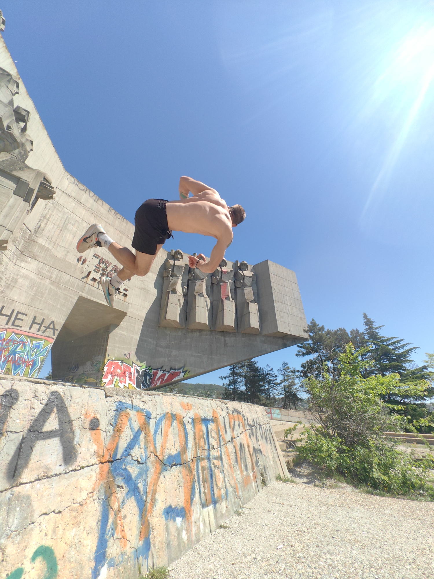 A shirtless man wearing black shorts and athletic shoes is airborne, positioned horizontally in mid-jump. He is in front of a large, concrete monument with Soviet-era blocky designs and some trees. The monument is covered with graffiti. Below the monument is a lower concrete wall, also covered in graffiti. The area around the wall is dirt and gravel. The sky is bright blue with the sun at the right. The location is the Buzludzha Monument in Buzludzha, Bulgaria.