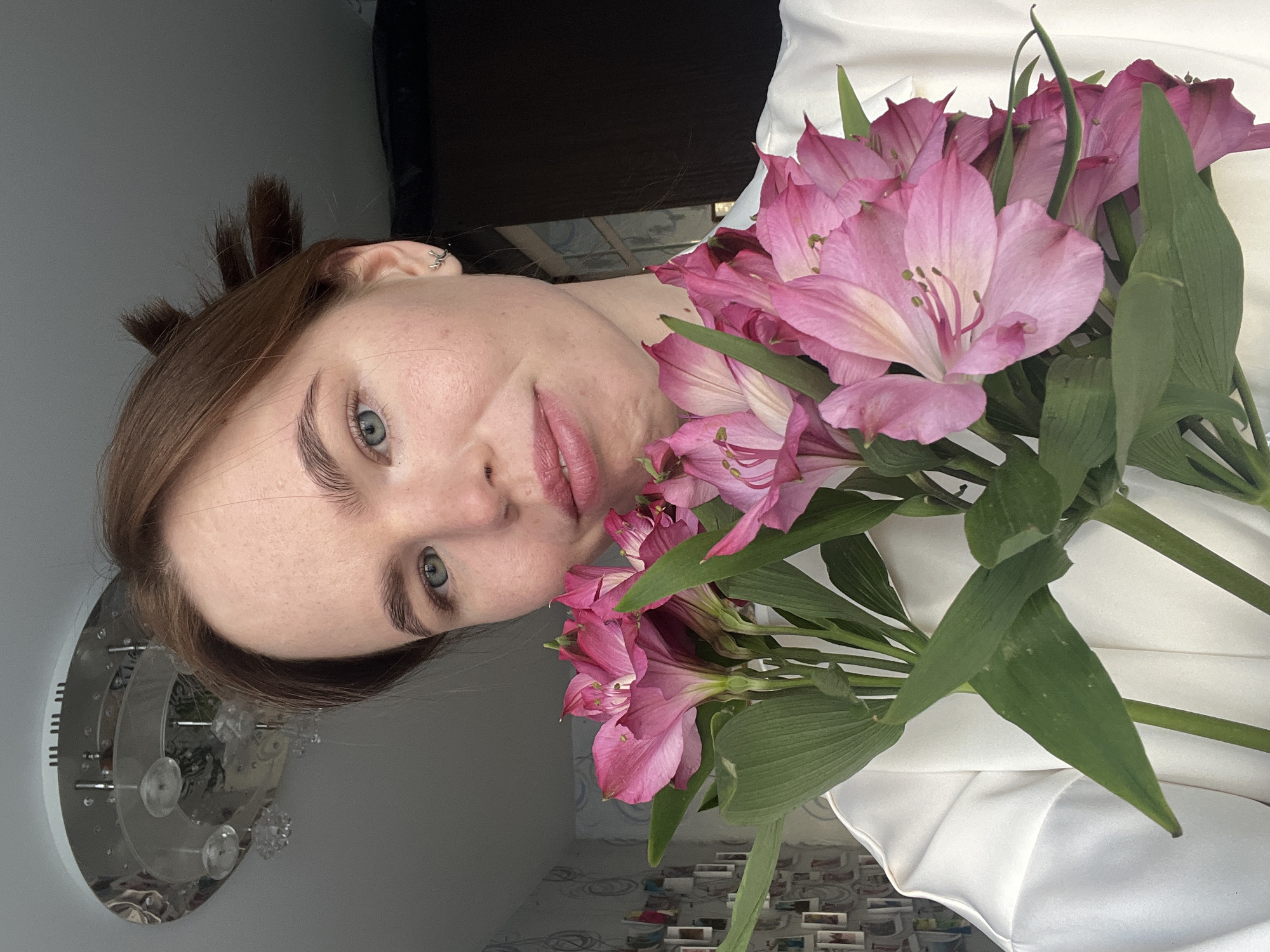 A female individual, appearing in her 20s to early 30s, is depicted in a close-up, chest-up, indoor photograph. She has fair skin, light blue-grey eyes, natural eyebrows, and brown hair styled with visible updos on the right side of her head. A small, metallic, curved earring is present in her right ear. She is wearing a white garment, likely a shirt or blouse. Her lips are slightly parted, exhibiting a light pink coloration. She holds a bouquet of approximately seven to ten vibrant pink Alstroemeria flowers with prominent green stems and leaves, positioned centrally in front of her upper torso, partially obscuring her lower face. The background consists of a white ceiling featuring a circular, chrome and glass ceiling light fixture with decorative elements in the upper left quadrant. To the subject's left, a wall with light-colored, patterned wallpaper is visible, adorned with a collage of multiple small framed photographs or postcards. The area to the subject's right is darker and less defined, suggesting a further recessed space or dark furniture, with a partial view of a window through a doorway frame. The scene is located in an indoor setting within the city of Chaikovsky, Russia.