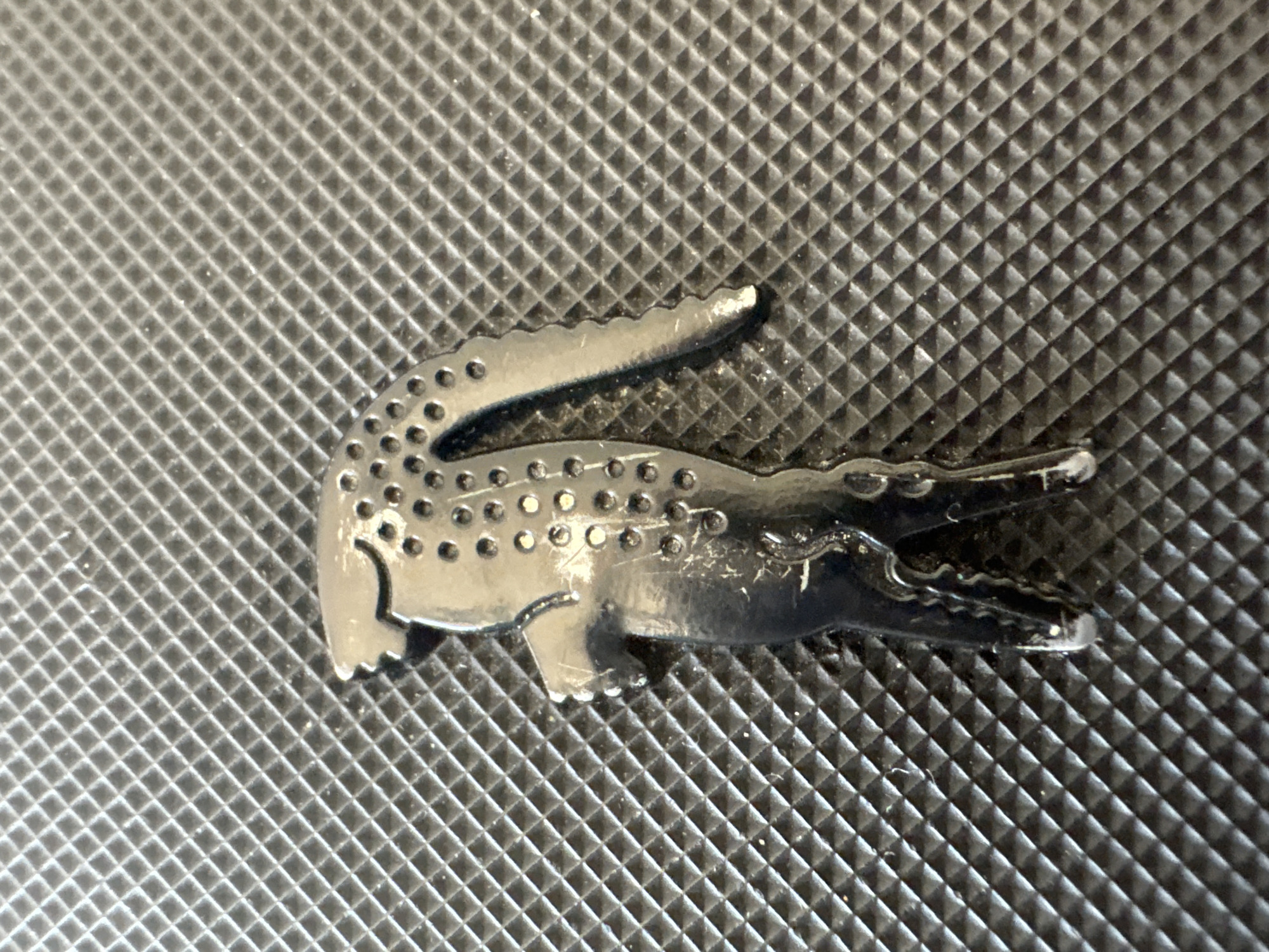 A close-up, high-angle shot features a metallic crocodile emblem, widely recognized as the Lacoste brand logo, centered against a dark, textured background. The emblem, rendered in a silver-grey metallic finish, depicts a crocodile with its head facing right and its mouth agape, revealing small, sharp teeth. Its back is adorned with numerous small, raised circular dots, simulating scales. Visible signs of wear, including minor scratches and scuff marks, are present across the emblem's surface, particularly around the mouth and body.
The background consists of a uniform, dark-colored (likely black or dark grey) textured material. This texture is a grid-like pattern of raised, small square or diamond shapes, creating a consistent three-dimensional surface. The lighting highlights the metallic sheen of the emblem and the contours of the background texture. There is no discernible activity taking place, nor are there any people or visible text in the image. The setting appears to be a static display of an object, possibly a part of a larger item like a bag, wallet, or accessory. No specific time of day, weather, or location cues are apparent due to the extreme close-up nature of the shot.