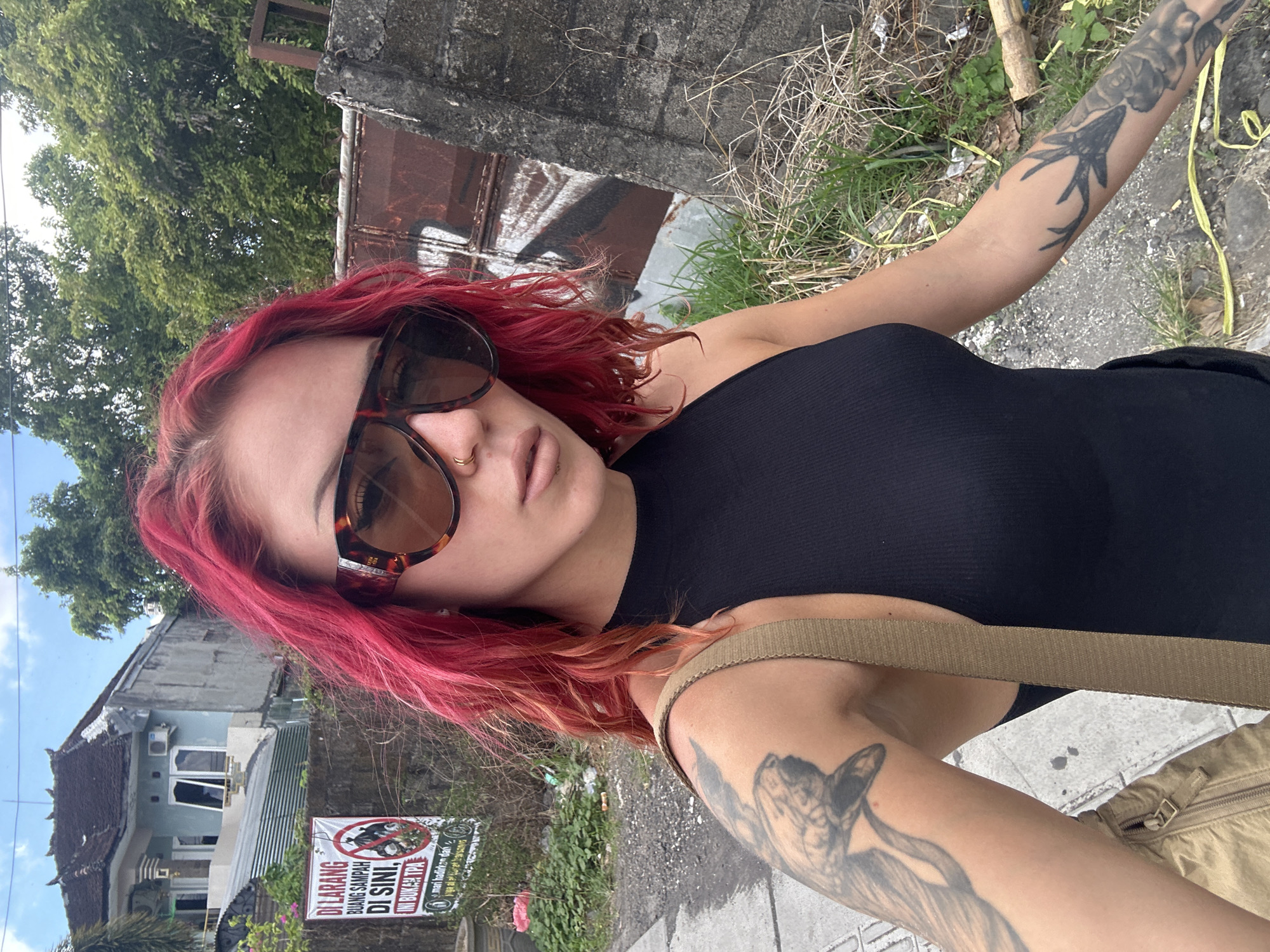 A woman with red hair, wearing a black top and brown sunglasses, stands on a sidewalk in North Kuta, Indonesia. She has tattoos on both arms and is wearing a brown shoulder bag. Behind her is a street with a house and a sign in Indonesian that reads "DILARANG BUANG SAMPAH DI SINI" (No littering here). The environment includes a concrete wall with some rusty metal attached and some vegetation. The overall scene suggests a casual, urban setting in a tropical location.