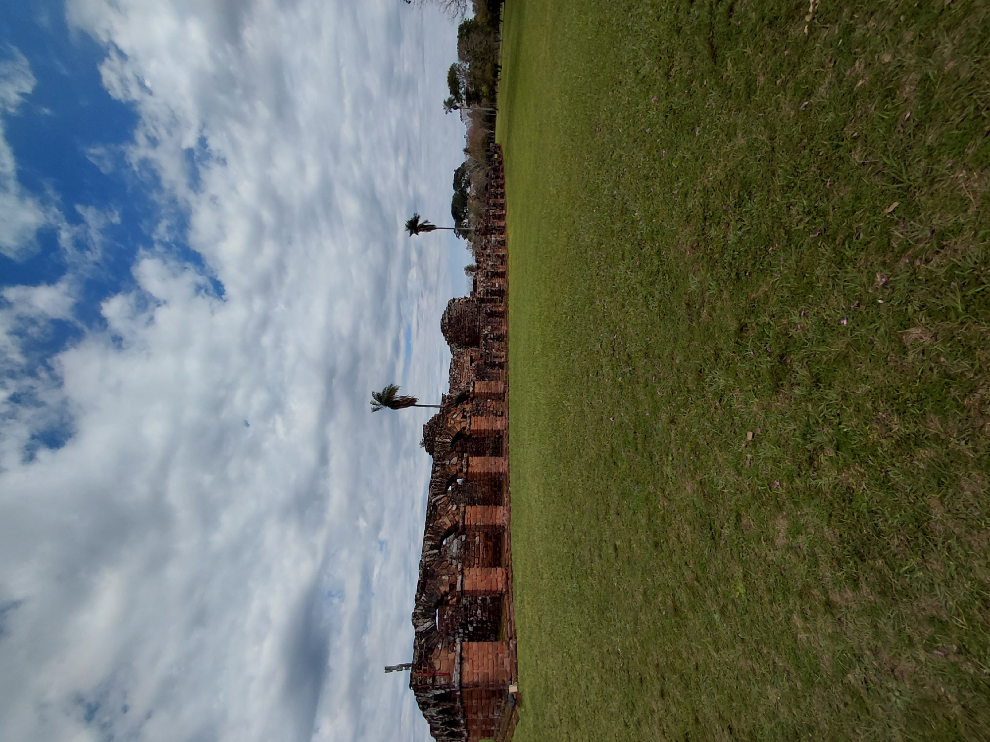 The image captures a panoramic view of the Ruins of the Holy Trinity of Parana, a UNESCO World Heritage site located in the city of Trinidad, Paraguay. The foreground is dominated by a vast expanse of lush green grass, manicured and evenly spread, with scattered fallen leaves and small purple flowers. The middle ground features the impressive ruins of a Jesuit mission, characterized by weathered, reddish-brown brick walls and arched windows. These structures appear ancient, with visible signs of decay and erosion, yet retain a sense of their former grandeur. Two tall, slender palm trees stand prominently within the ruins, adding a tropical element to the scene. In the background, a dense line of trees forms a natural boundary, with more palm trees interspersed. The sky above is a dramatic mix of cloudy and clear blue patches, with fluffy white clouds casting shadows on the landscape below, suggesting dynamic weather conditions. The overall impression is one of historical significance set against a natural, open environment.