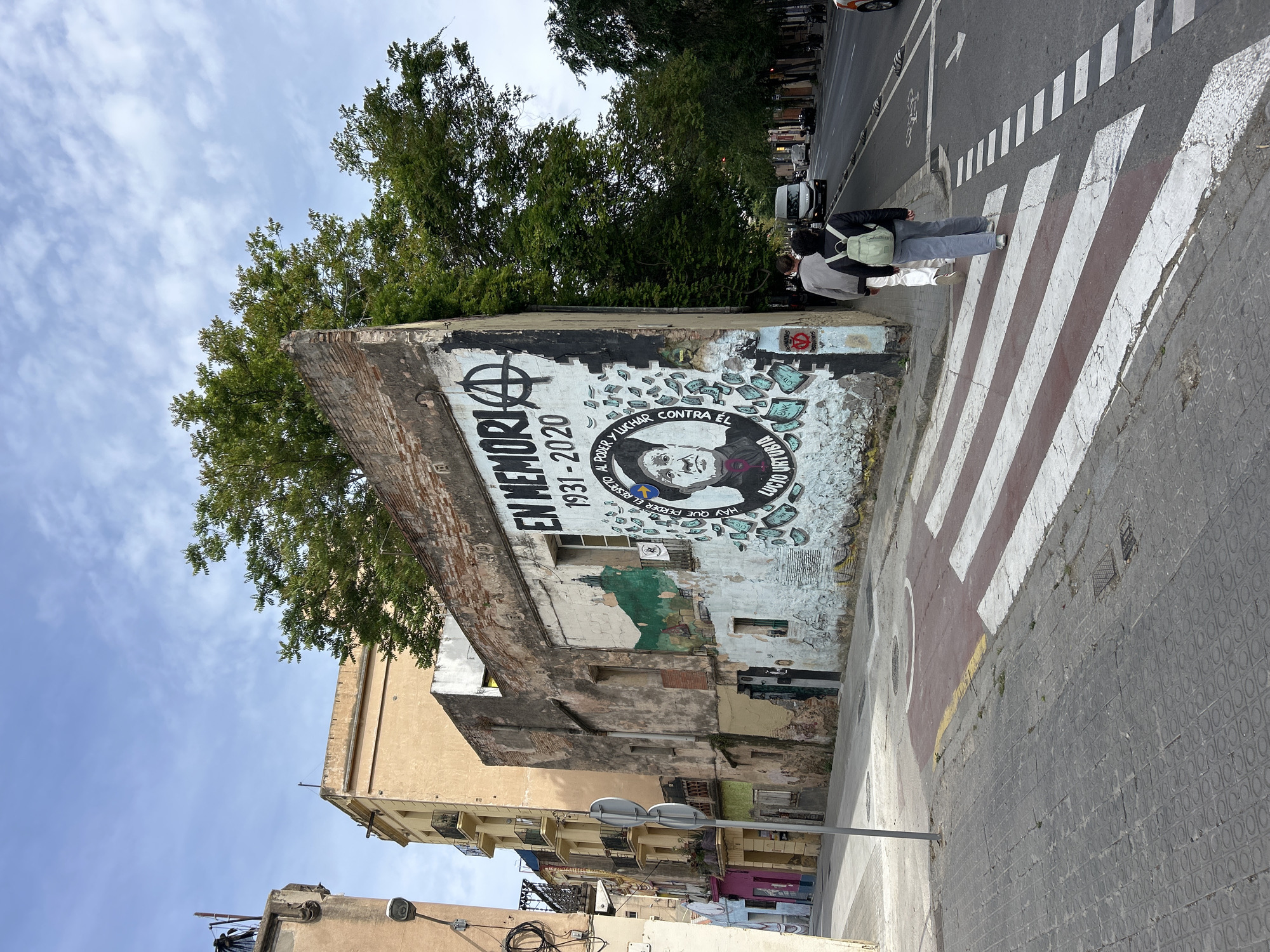 Here's a detailed description of the image:
The image captures an urban scene featuring a mural located in Barcelona, Spain. The mural is painted on the side of a dilapidated building, with part of the wall crumbling and revealing exposed brick. The artwork commemorates Lucio Urtubia, with the text "EN MEMORIA 1931-2020" and a portrait of Urtubia. An anarchy symbol is prominently displayed above the text. Additional text on the mural states, "HAY QUE PERDER EL RESPETO AL PODER Y LUCHAR CONTRA EL," translating to "We must lose respect for power and fight against it." Imagery of currency is also incorporated into the design.
Two individuals are walking across a pedestrian crosswalk in front of the mural. They are dressed casually.
The street scene includes a sidewalk, a crosswalk with alternating red and white stripes, and a street with vehicular traffic. Buildings of varying architectural styles line the street, with trees visible in the background. The sky is partly cloudy. A street sign is visible on the lower part of the image.