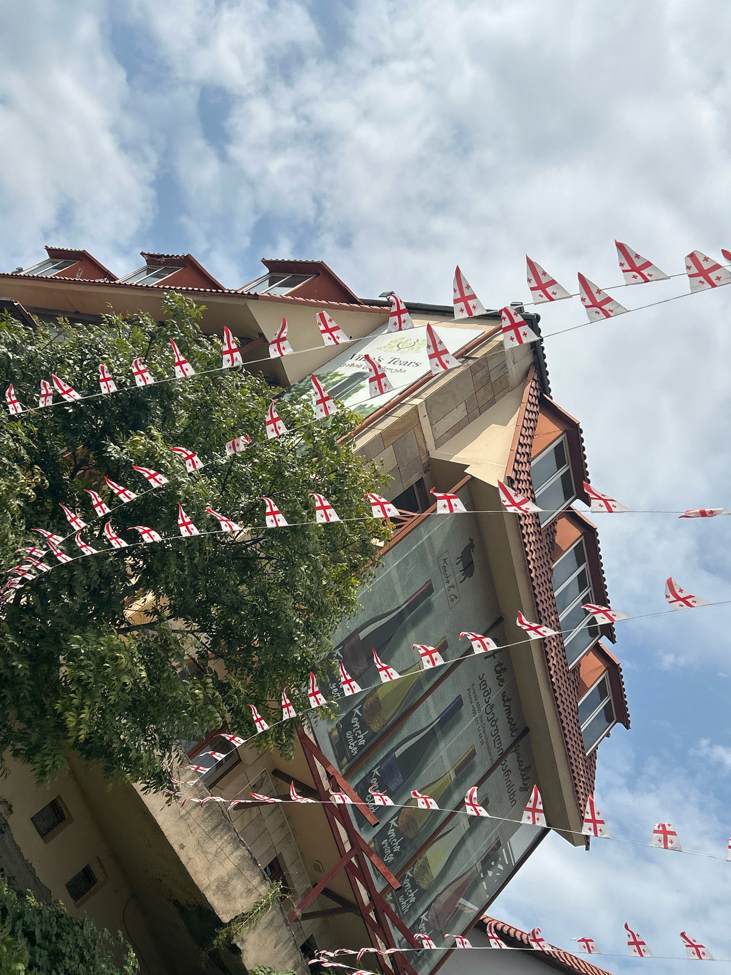 The image depicts a street view in the city of Sighnaghi, Georgia, adorned with strings of small triangular flags featuring the Georgian national flag design. The flags are strung across a building facade which appears to be a shop or restaurant, indicated by a large mural on the side of the building. The mural shows several wine bottles and text that appears to be in both English and Georgian. The building has a tiled roof and several windows. A large tree with green foliage is partially visible in the foreground, and the sky above is partly cloudy. The perspective of the image is tilted, giving a dynamic but slightly disorienting view of the scene. There are no people visible in the image.