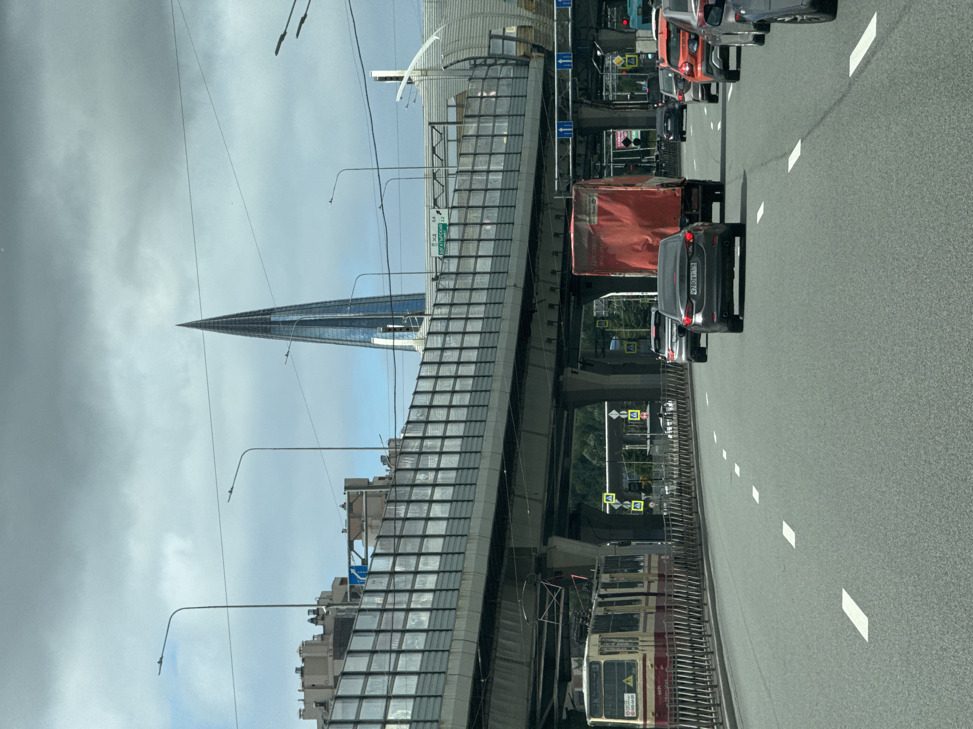 This is a medium-distance, eye-level shot of a busy highway in Saint Petersburg, Russia. A cloudy sky looms overhead, with a tall, slender skyscraper piercing the clouds in the distance. The highway itself is lined with vehicles, including a red truck, several cars, and an older model tram. An elevated highway structure with a transparent sound barrier runs overhead, and streetlights are visible along the roadside. The scene appears to be a typical urban commute on a slightly overcast day.
