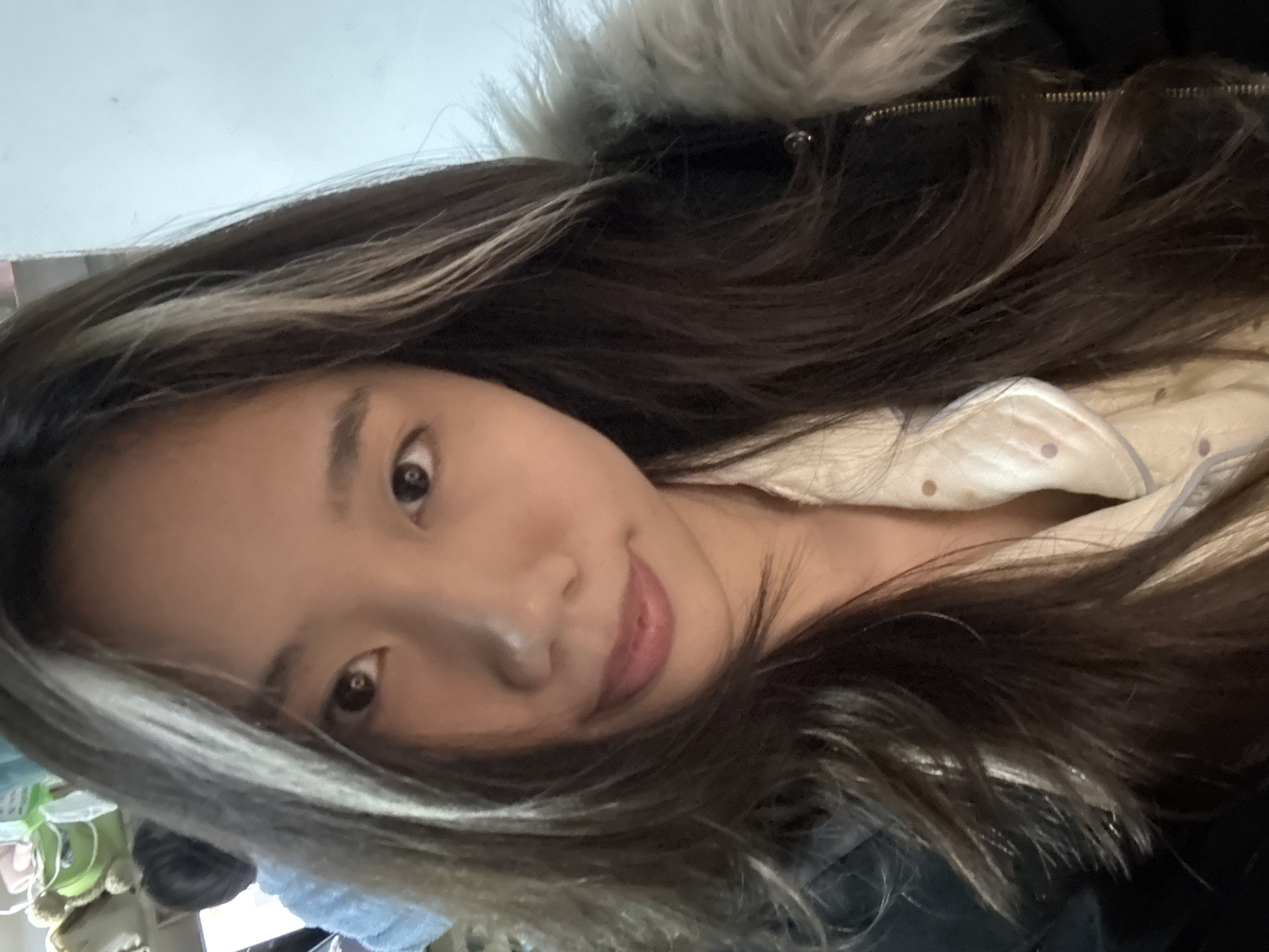 A close-up indoor portrait features a young East Asian female in a residential setting within Nan'an District, China. The primary subject has long, dark brown hair with distinct light blonde/grey streaks on the viewer's left side, framing her face. She has dark eyes and a subtle smile on her lips. Her attire consists of a light-colored, polka-dotted collared shirt beneath a dark-colored jacket, which features a prominent grey and white faux fur collar. The subject is looking directly at the camera. In the out-of-focus background to the left, the head of a second individual, possibly a child, is partially visible, wearing a light blue, textured garment. Behind this individual, a light green container and several brown plush toys are discernible. The background to the right consists of a plain, light blue wall.